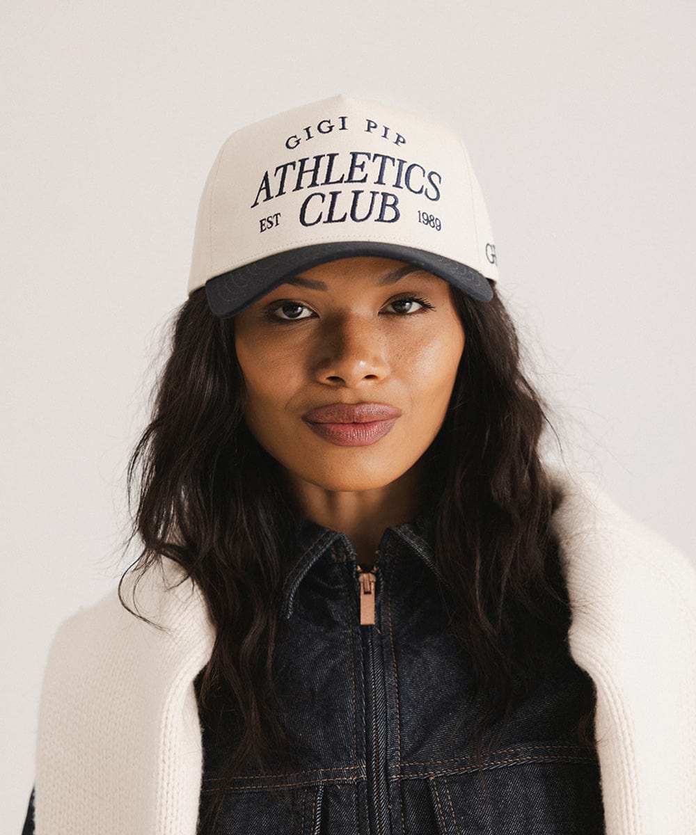 Woman wearing a Navy Trucker hat with 'Gigi Pip Athletics Club' text and a denim jacket against a plain background #color_navy