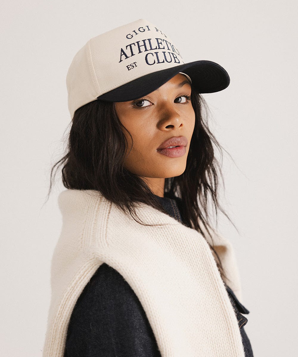 Woman wearing a Navy Trucker hat with 'Gigi Pip Athletics Club' text and a denim jacket, posing sideways against a plain background #color_navy