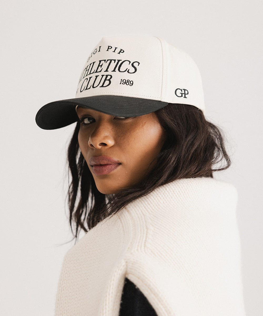Woman wearing a cream and black Trucker hat with 'Gigi Pip Athletics Club' text and a white sweater, posing sideways against a plain background #color_cream - black