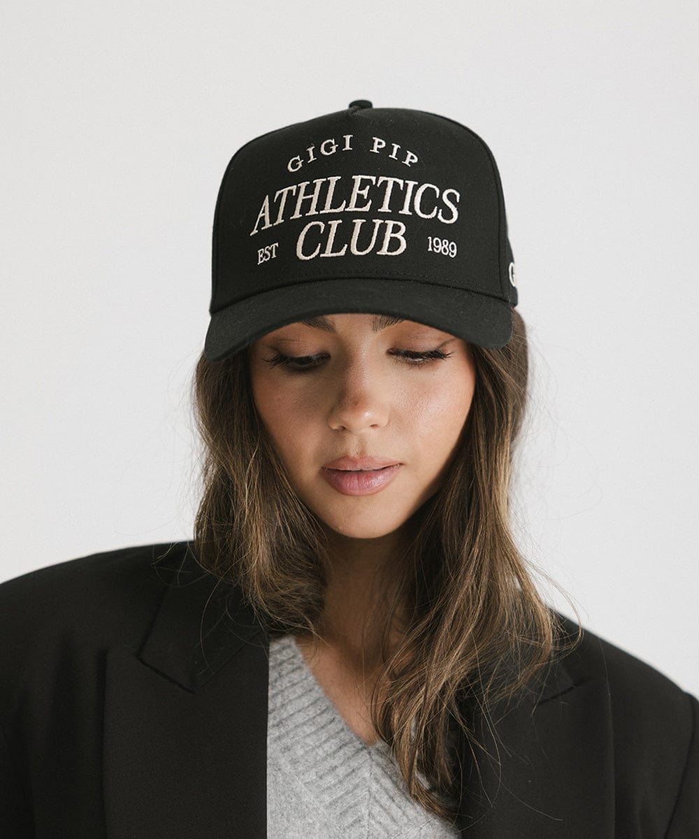 Woman wearing a Black Trucker hat with 'Gigi Pip Athletics Club' text and a black blazer against a plain background #color_black