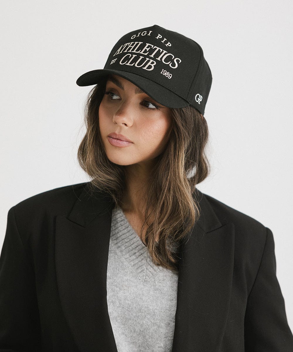 Woman wearing a Black Trucker hat with 'Gigi Pip Athletics Club' text and a grey top with a black blazer against a plain background #color_black