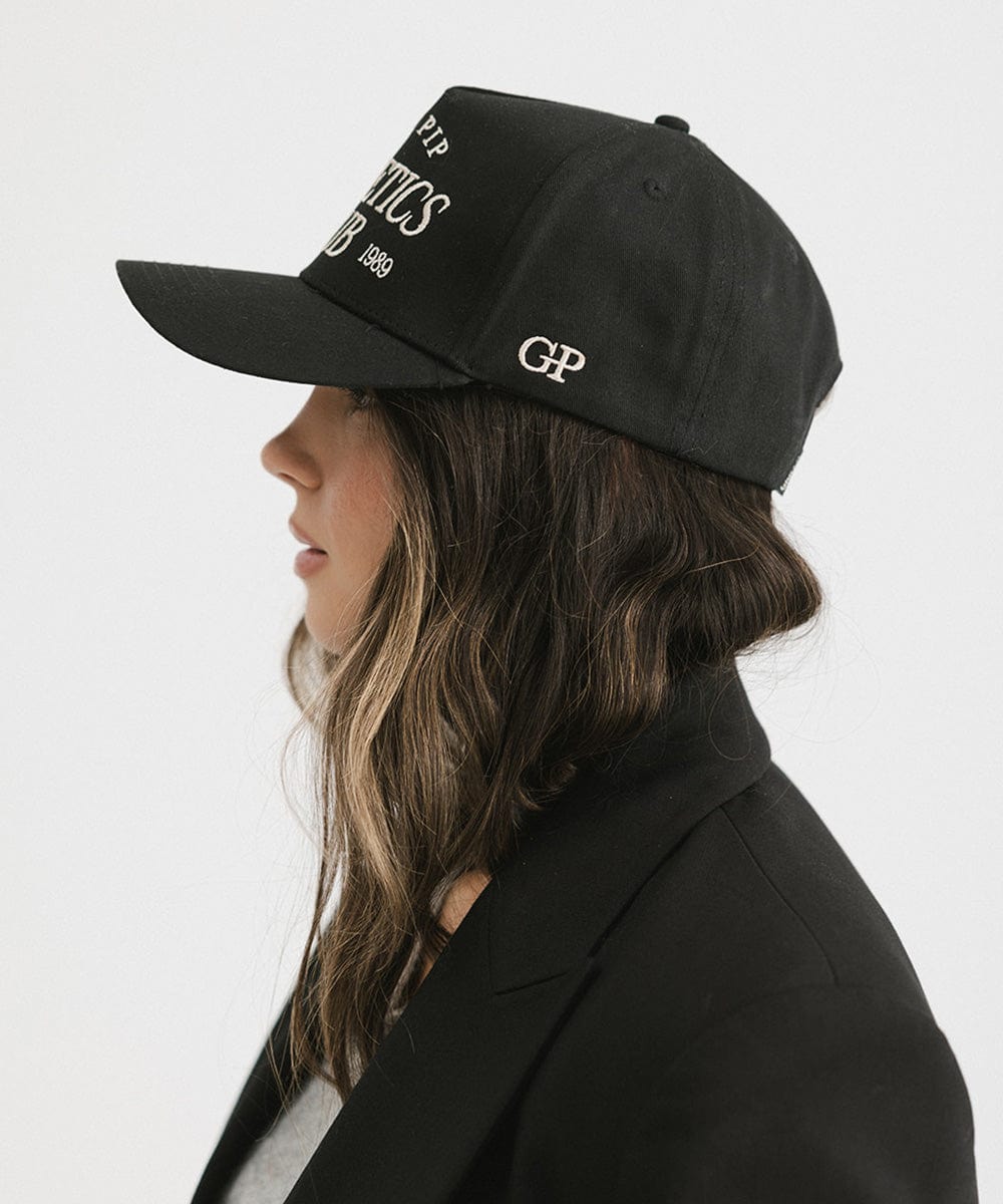 Woman wearing a Black Trucker hat and a black blazer, facing sideways against a plain background #color_black