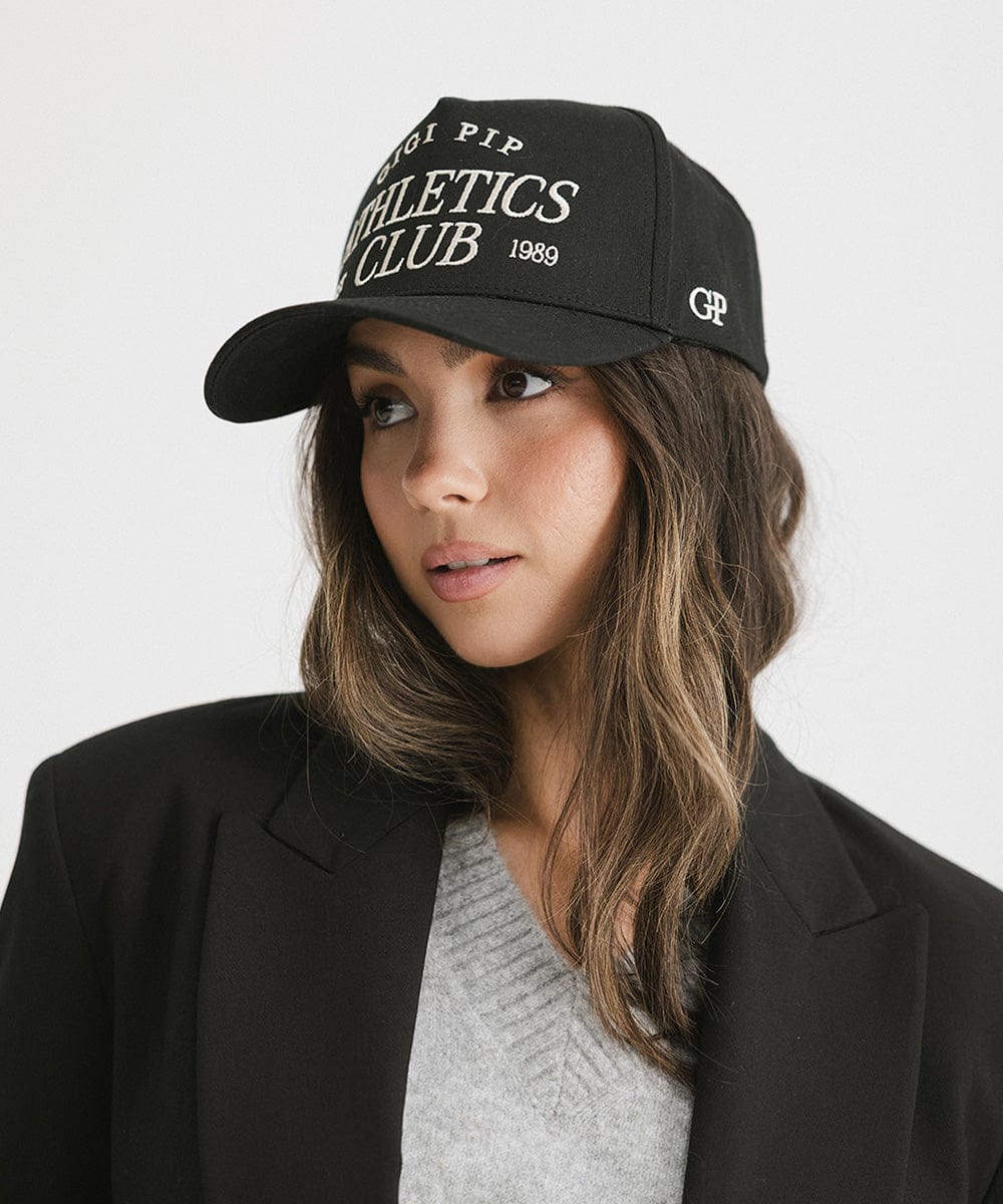 Woman wearing a Black Trucker hat with 'Gigi Pip Athletics Club' text and a black blazer, posing against a plain background #color_black