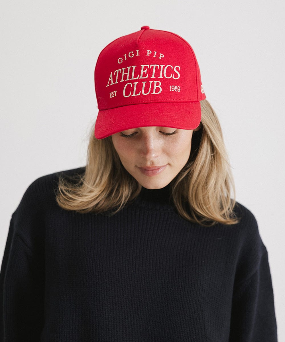 Woman wearing a Red Trucker hat with 'Gigi Pip Athletics Club' text and a dark blue sweater, posing against a plain background #color_red