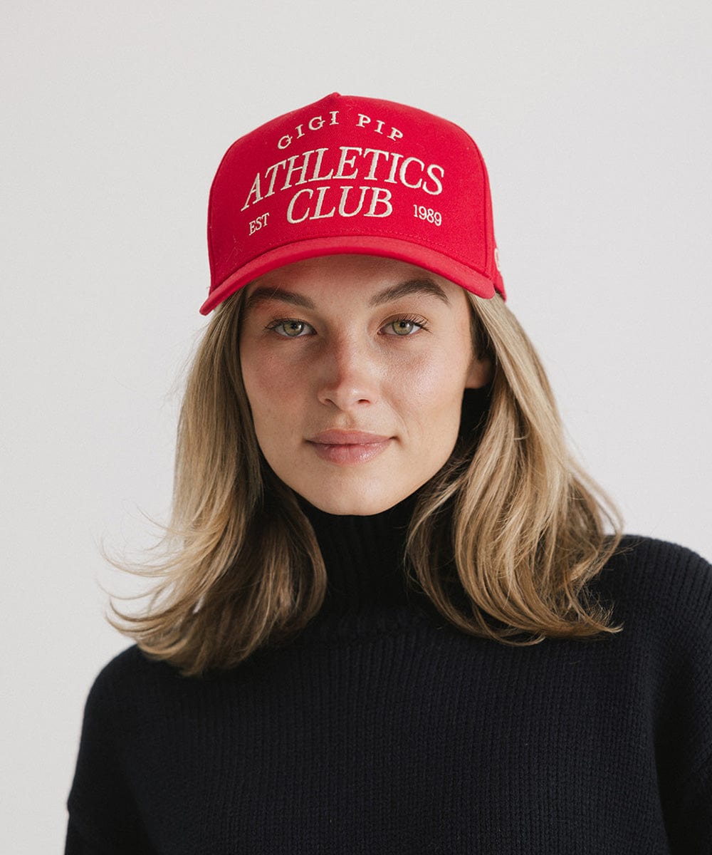 Woman wearing a Red Trucker hat with 'Gigi Pip Athletics Club' text and a dark blue sweater against a plain background #color_red