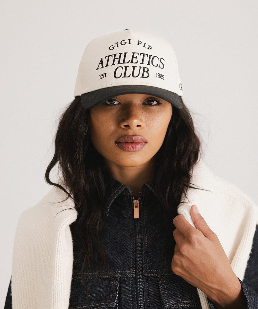 Woman wearing a cream and black Trucker hat with 'Gigi Pip Athletics Club' text and a denim jacket against a plain background #color_cream - black