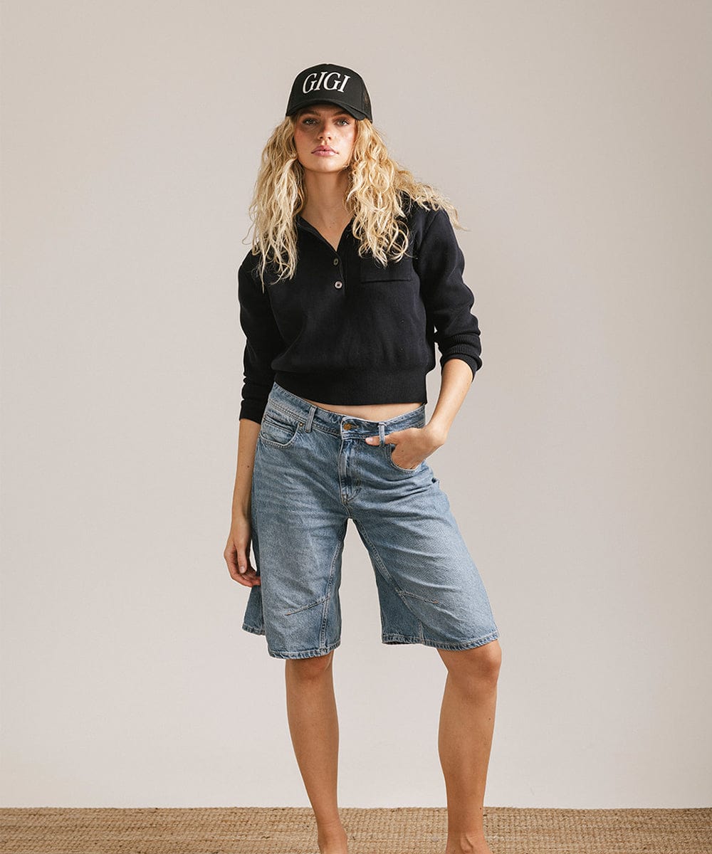 Woman wearing a black cap with 'GIGI' text, a black shirt and denim shorts against a plain background #color_black
