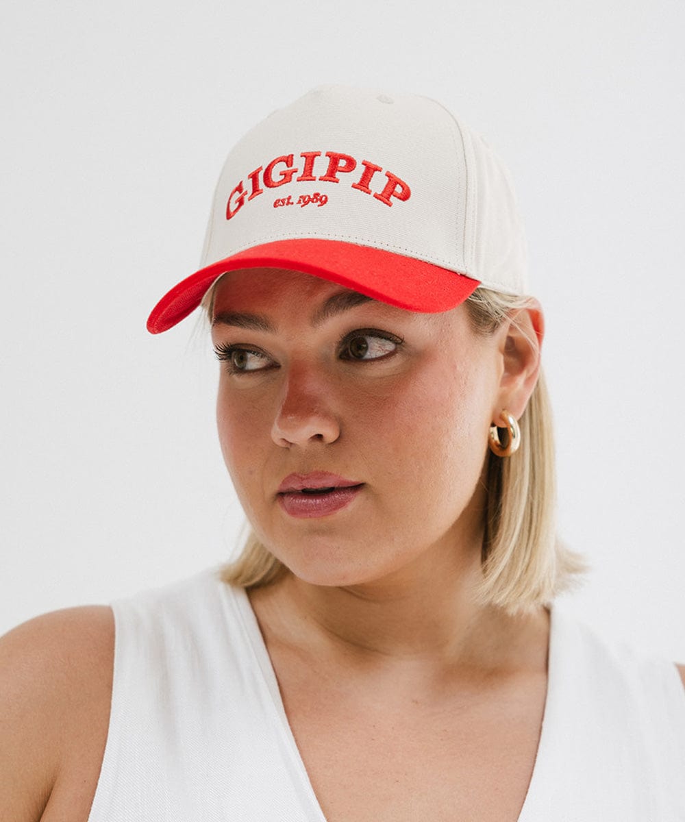 Woman wearing a cream and red trucker hat with 'GIGI PIP' embroidered in red on the front and a white top against a plain background #color_cream - red