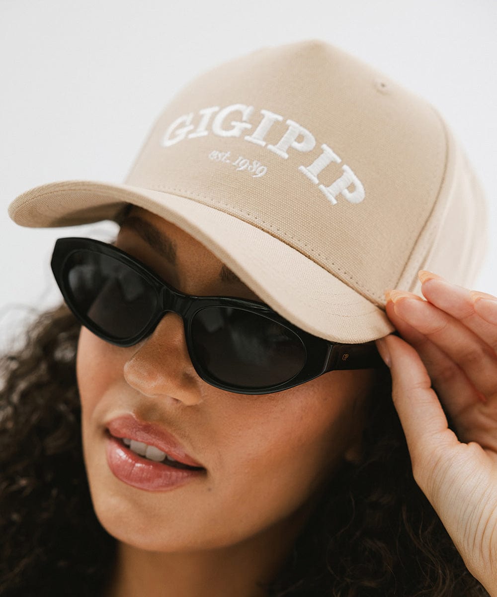 Woman wearing a tan trucker hat with 'GIGI PIP' embroidered in white on the front and black sunglasses against a plain background #color_tan