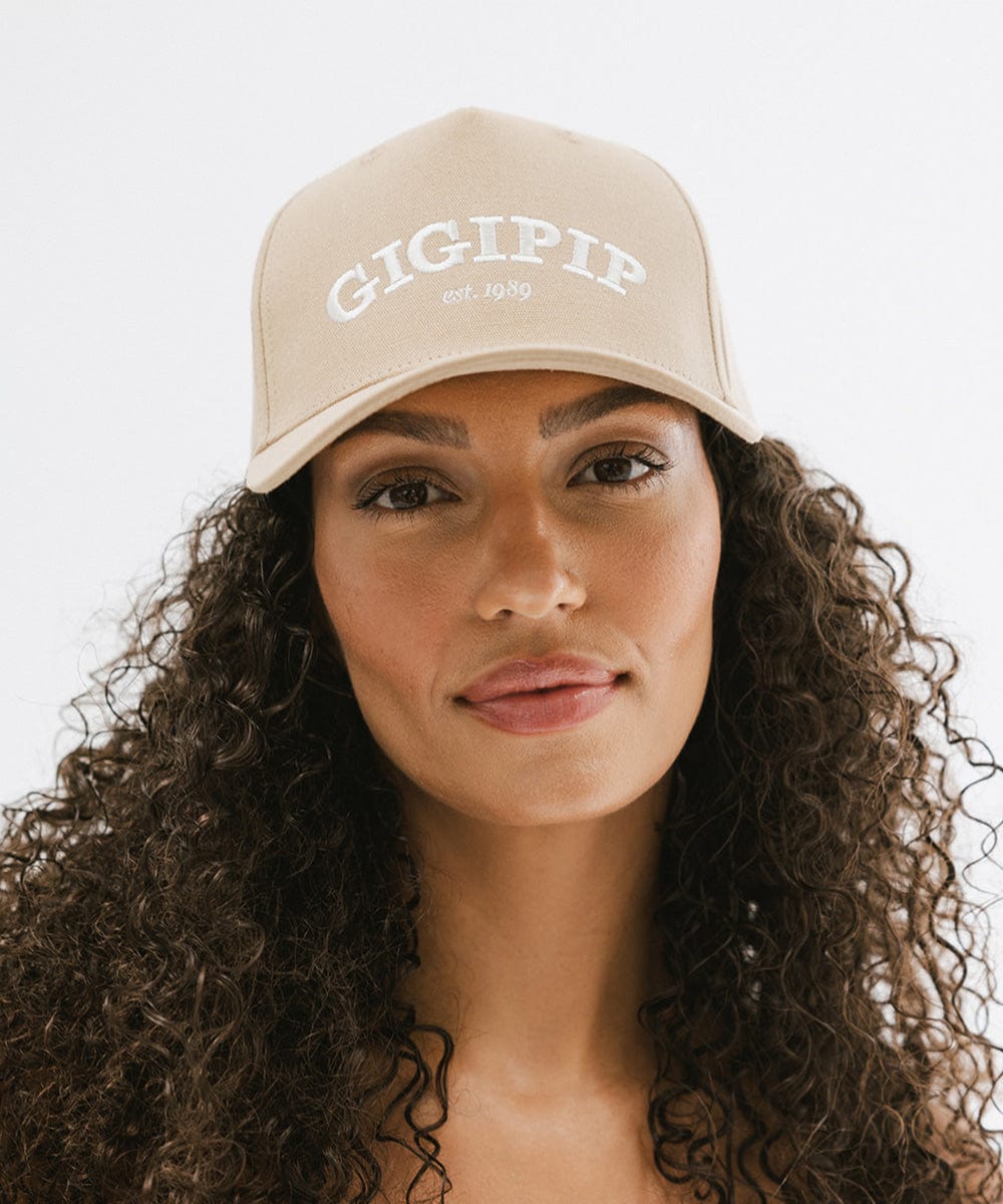 Woman wearing a tan trucker hat with 'GIGI PIP' embroidered in white on the front against a plain background #color_tan