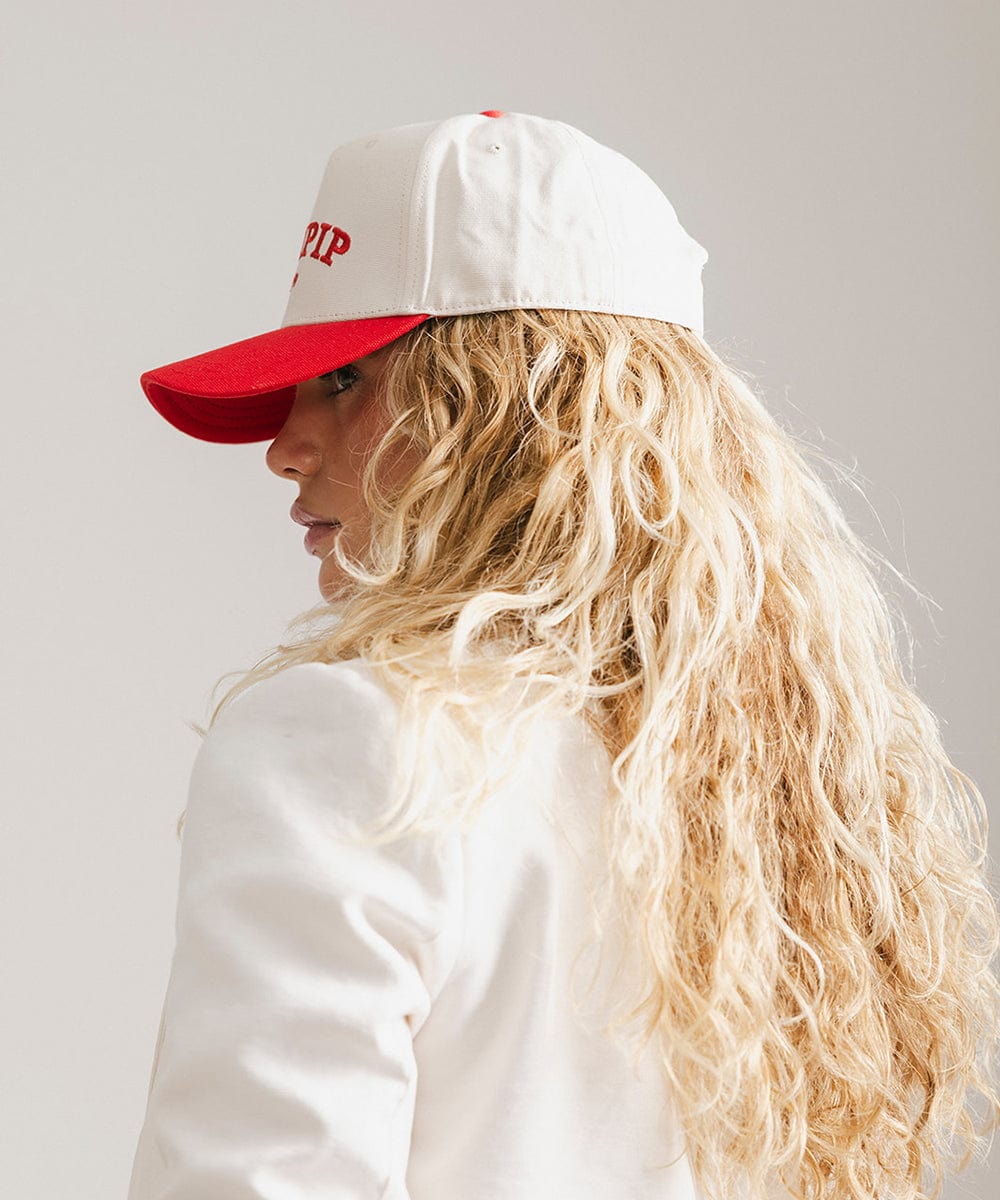 Woman wearing a cream and red trucker hat with 'GIGI PIP' embroidered in red on the front and a white coat, facing sideways against a plain background #color_cream - red