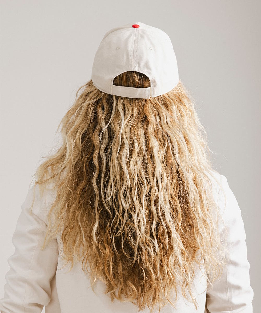 Woman with long hair wearing a cream and red trucker hat and a white coat against a plain background #color_cream - red