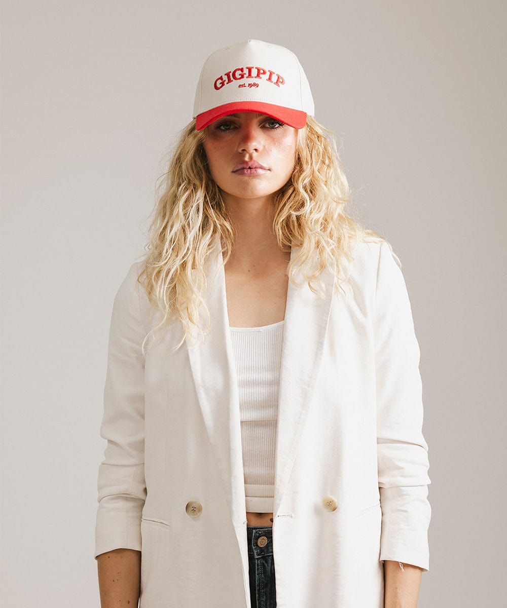 Woman wearing a cream and red trucker hat with 'GIGI PIP' embroidered in red on the front and a white coat against a plain background #color_cream - red