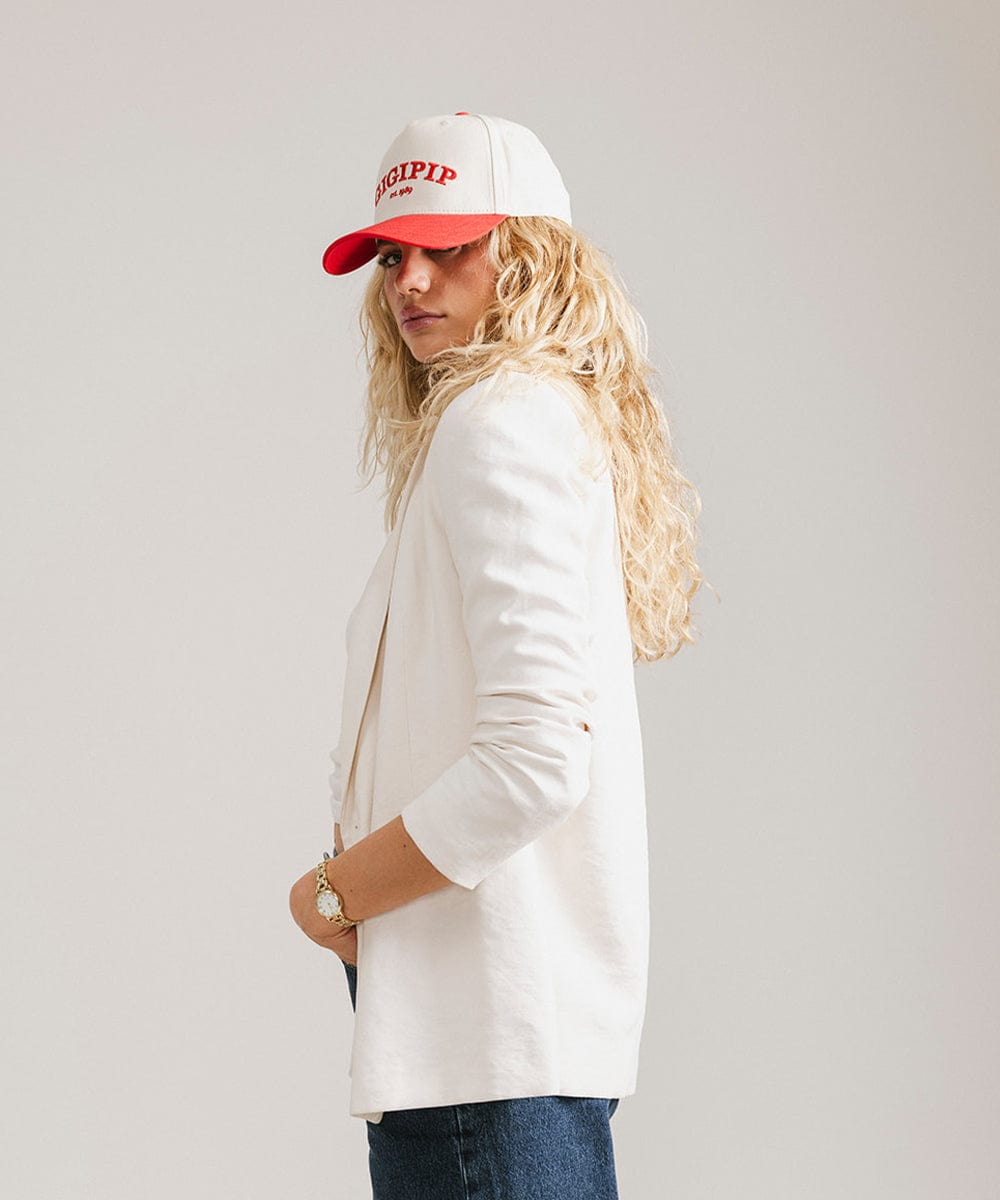 Woman wearing a cream and red trucker hat with 'GIGI PIP' embroidered in red on the front and a white coat, posing against a plain background #color_cream - red