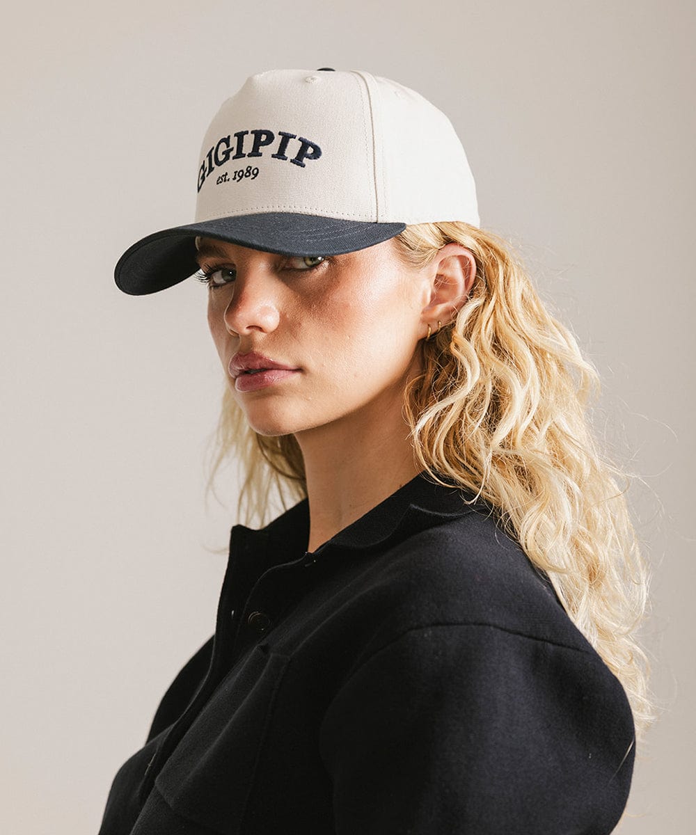 Woman wearing a cream and navy trucker hat with 'GIGI PIP' embroidered in navy on the front and a black shirt, posing sideways against a plain background #color_cream - navy