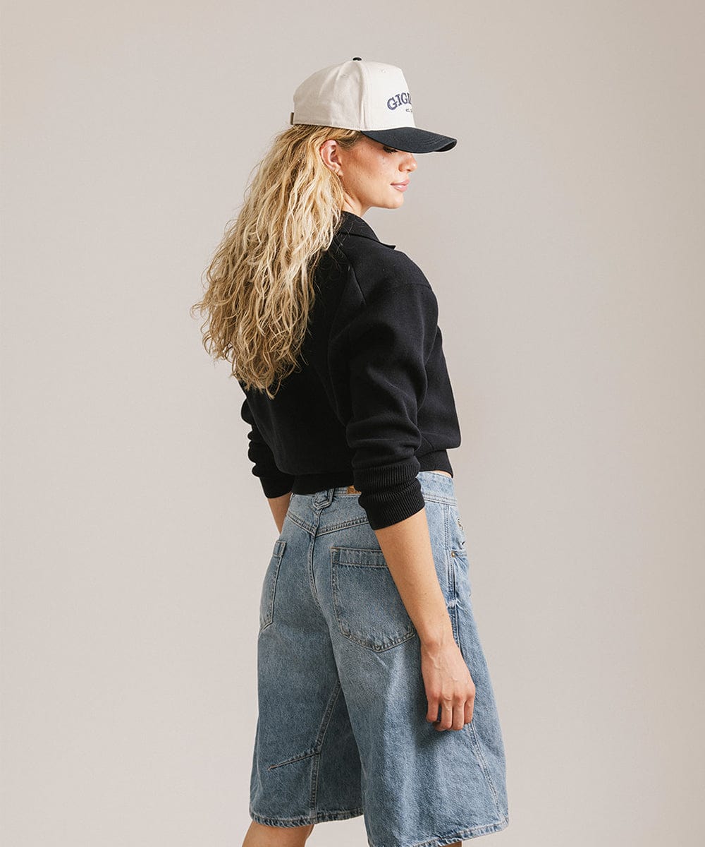 Woman wearing a cream and navy trucker hat, a black sweater and denim shorts, posing sideways against a plain background #color_cream - navy