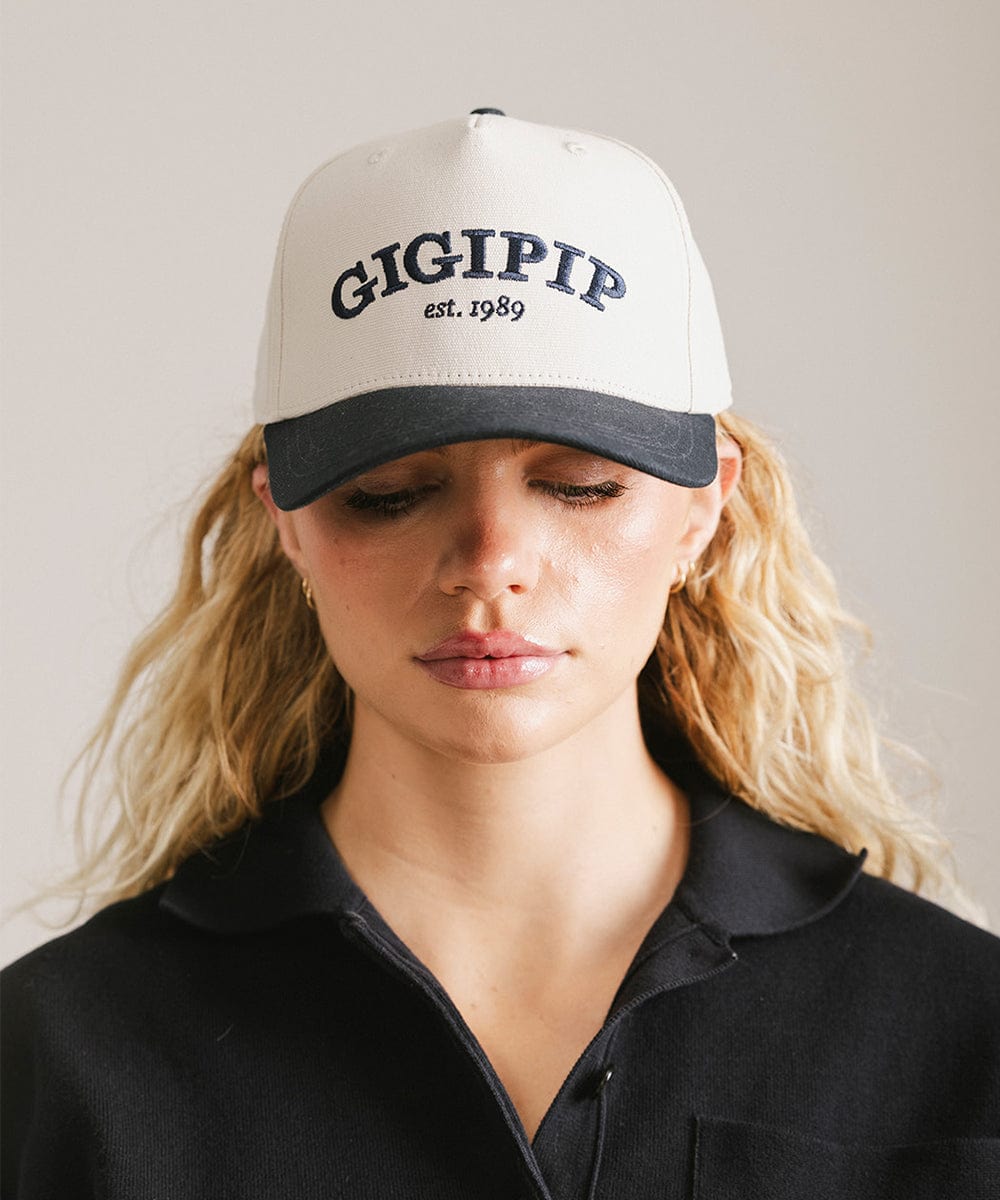 Woman wearing a cream and navy trucker hat with 'GIGI PIP' embroidered in navy on the front and a black shirt against a plain background #color_cream - navy