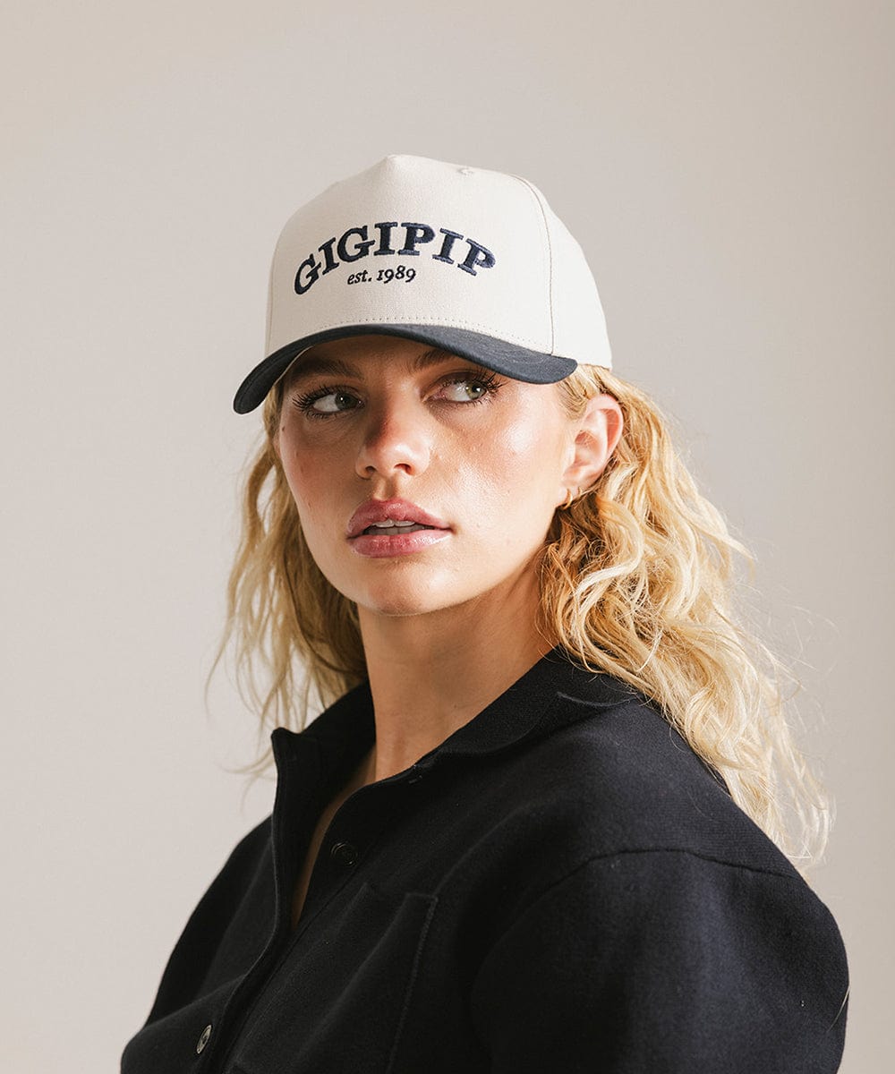 Woman wearing a cream and navy trucker hat with 'GIGI PIP' embroidered in navy on the front and a black shirt, posing against a plain background #color_cream - navy