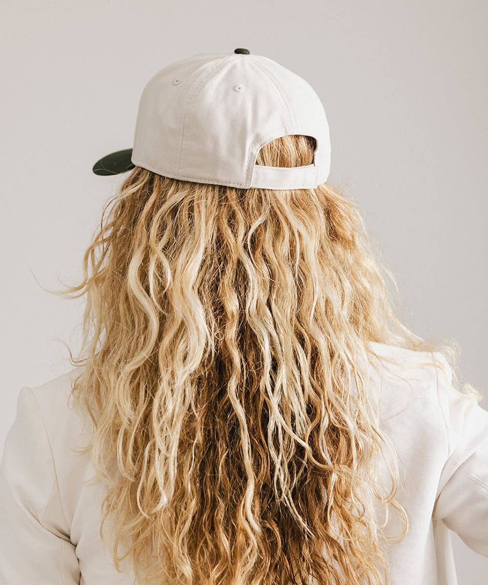 Woman with long hair wearing a cream and dark green trucker hat and a white coat against a plain background #color_cream - dark green