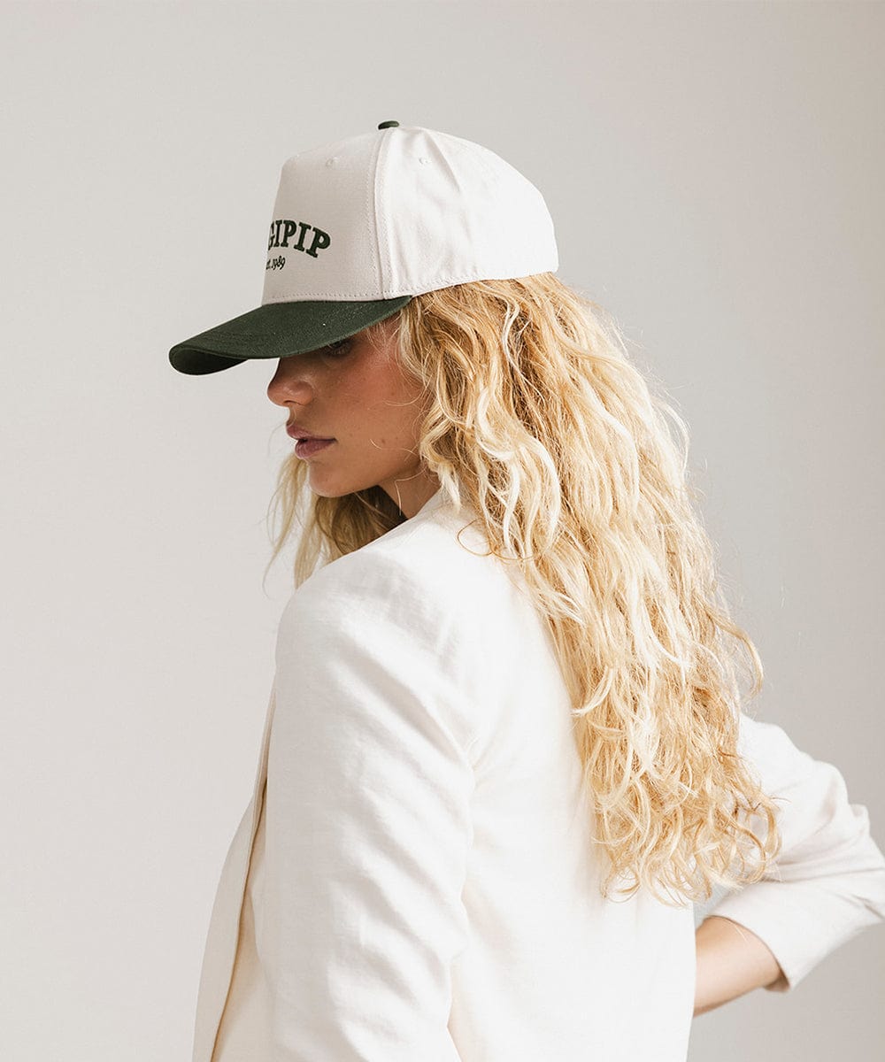Woman wearing a cream and dark green trucker hat with 'GIGI PIP' embroidered in dark green on the front and a white coat, posing sideways against a plain background #color_cream - dark green