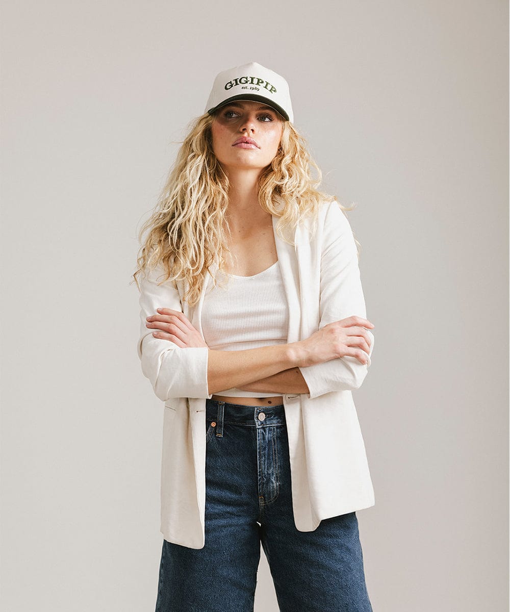 Woman wearing a cream and dark green trucker hat with 'GIGI PIP' embroidered in dark green on the front, a white top with a white coat and blue jeans against a plain background #color_cream - dark green