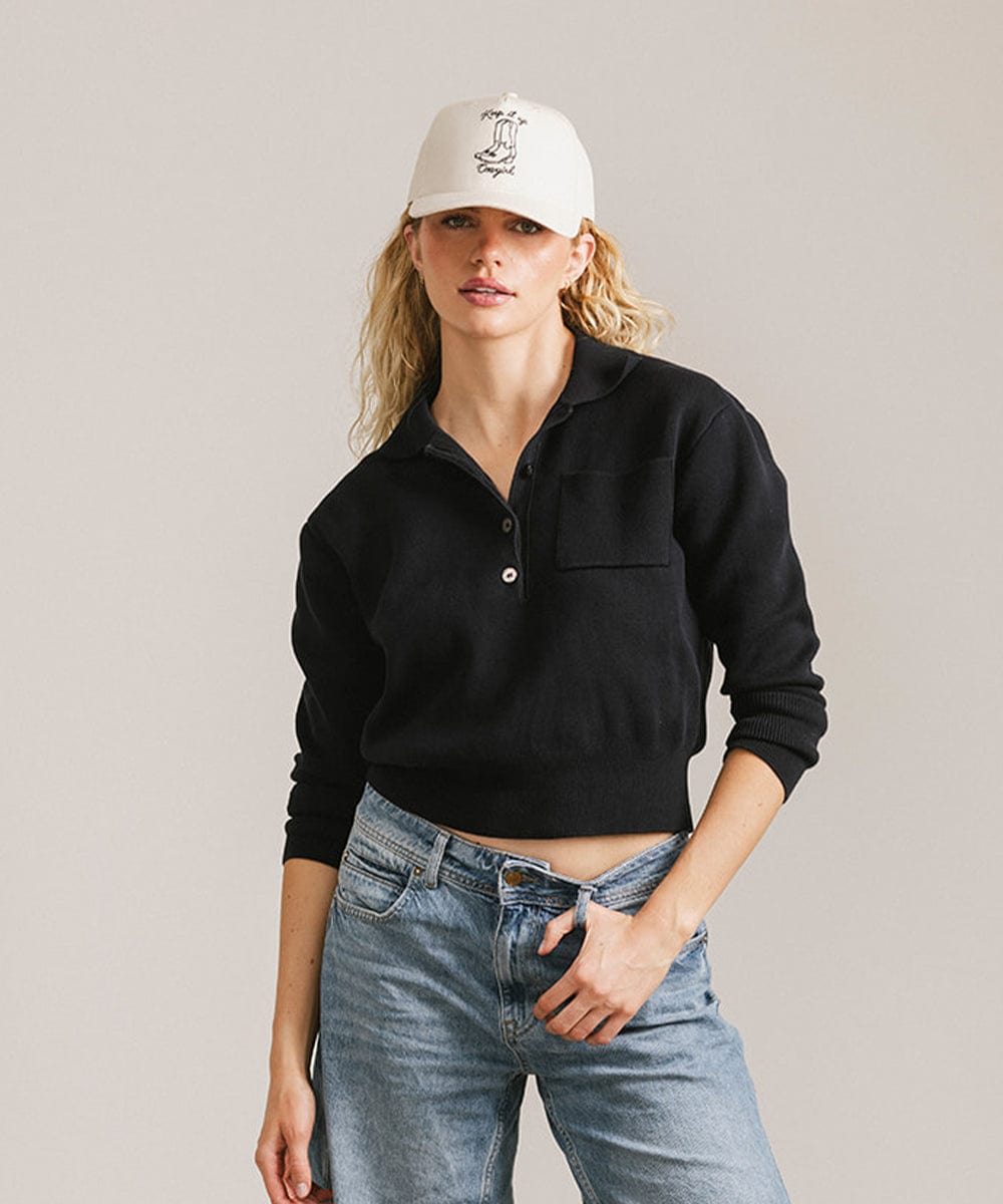 Woman wearing a cream cap with black text and boot design, a black shirt and blue jeans against a plain background #color_cream