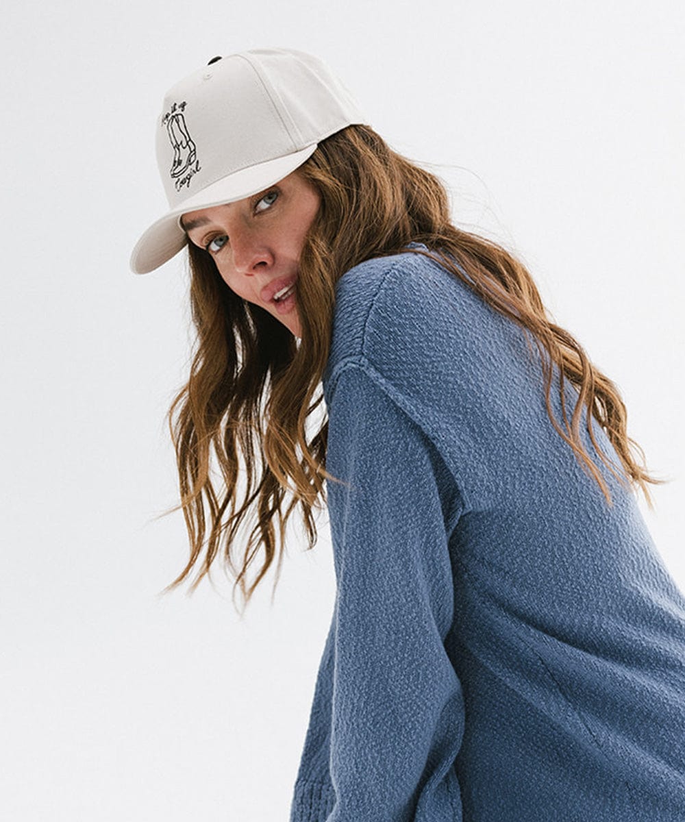 Woman wearing a cream cap with black text and boot design, and a blue sweater, posing sideways against a plain background #color_cream
