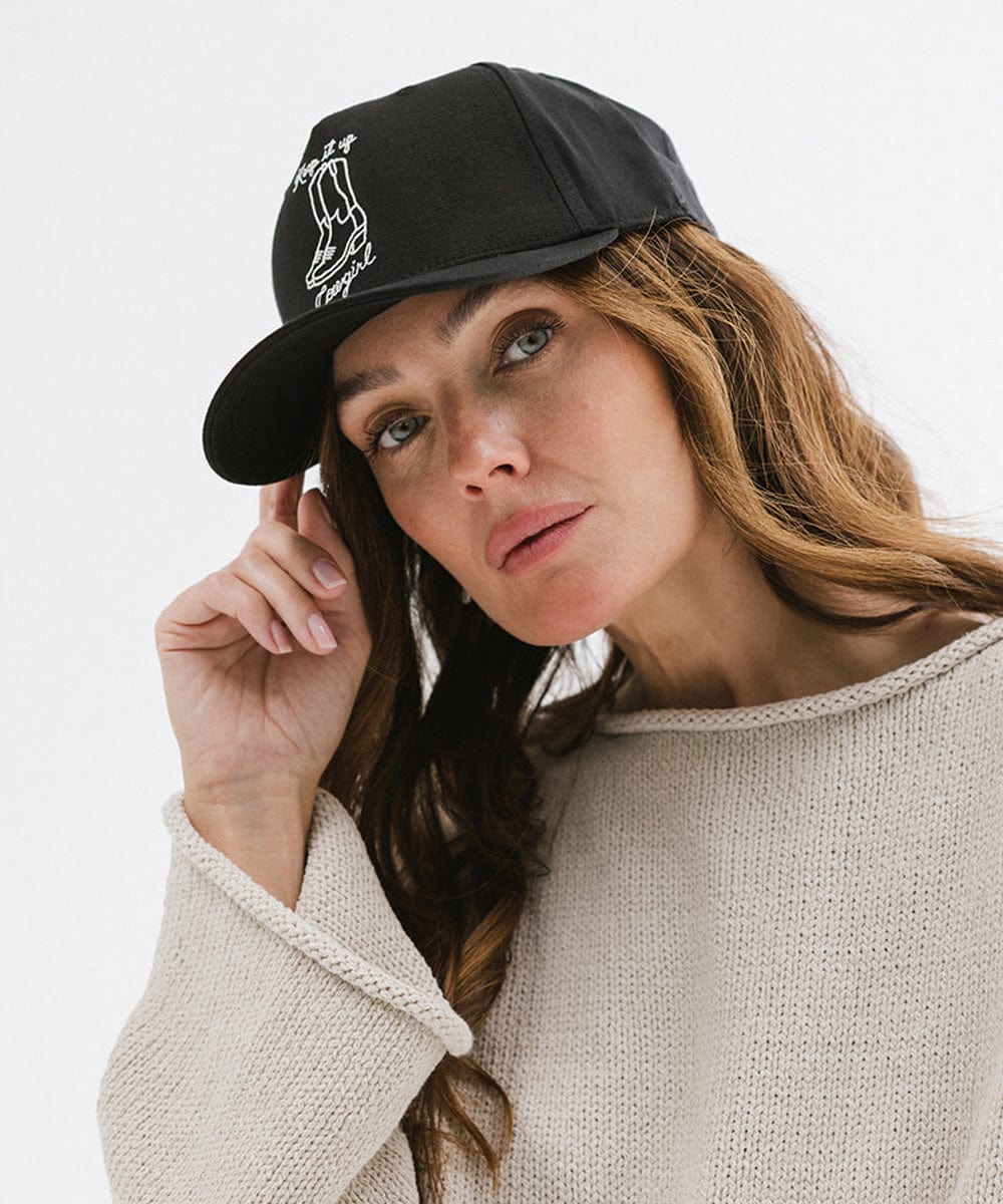 Woman wearing a black cap with white text and boot design and an off white t-shirt against a plain background #color_black