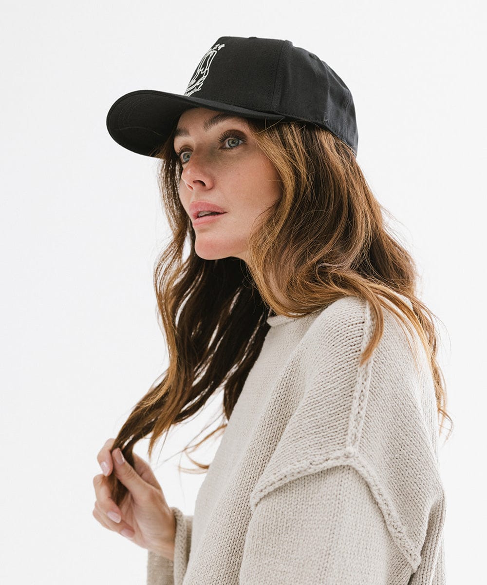 Woman wearing a black cap with white text and boot design and an off white t-shirt, posing sideways against a plain background #color_black