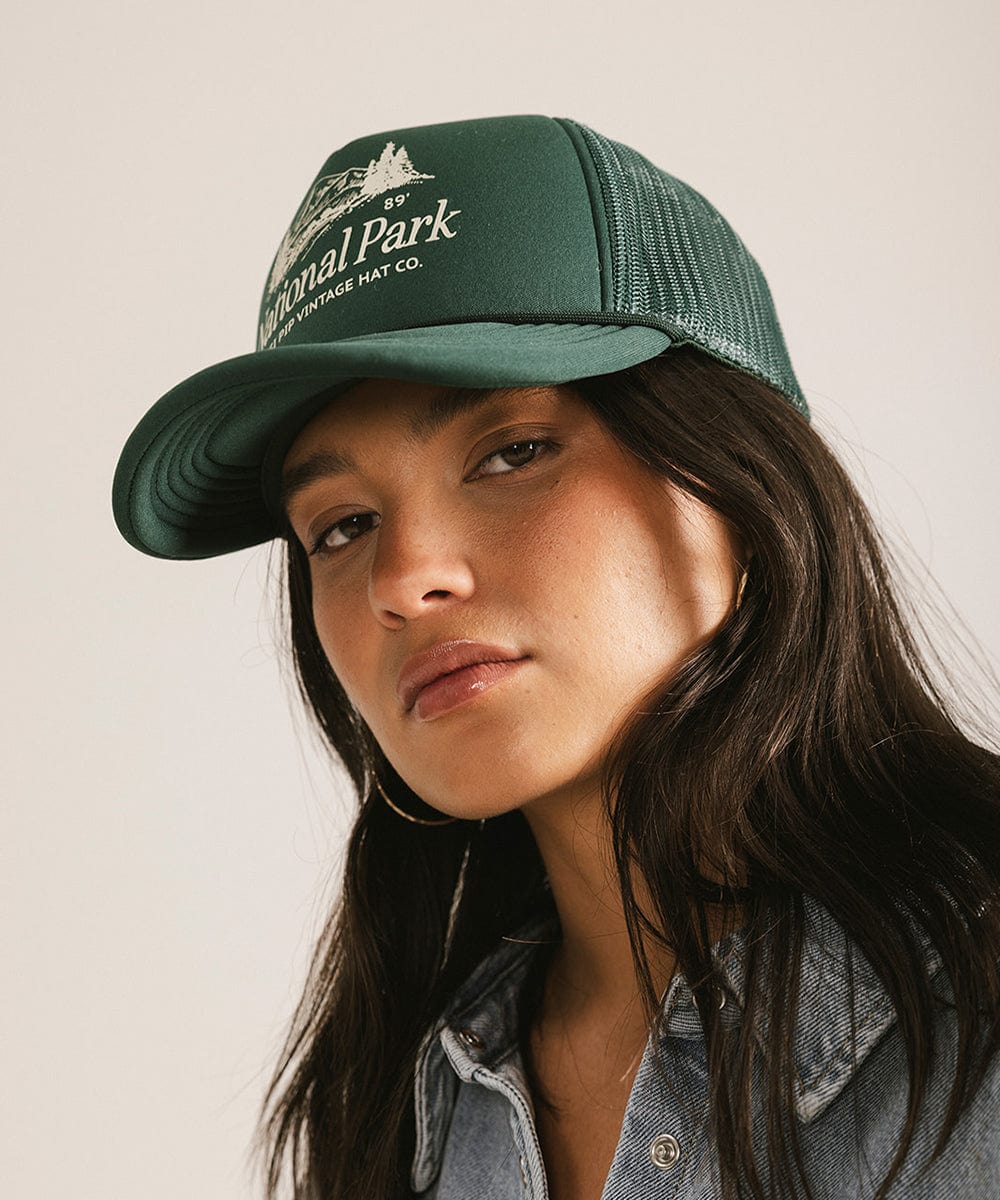 Woman wearing a forest green colored trucker hat with 'National Park' logo against a plain background #color_forest green