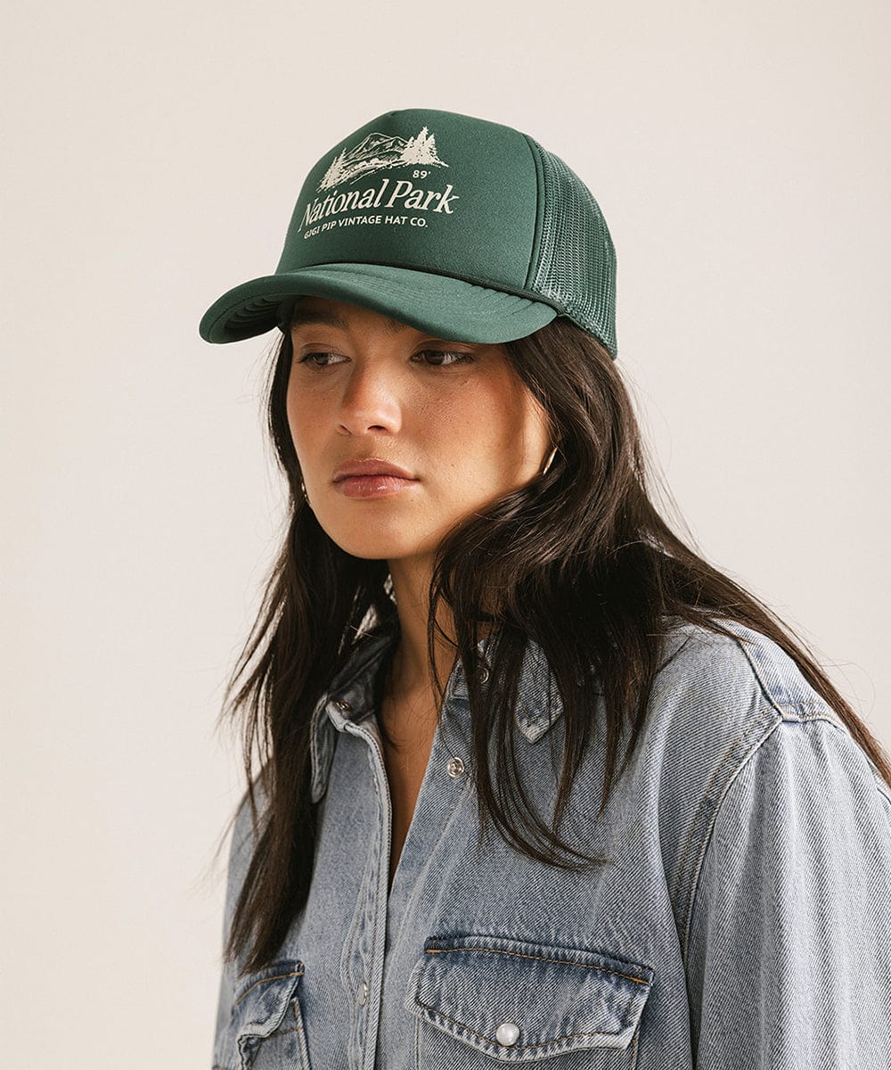 Woman wearing a forest green colored trucker hat with 'National Park' logo and a denim shirt against a plain background #color_forest green