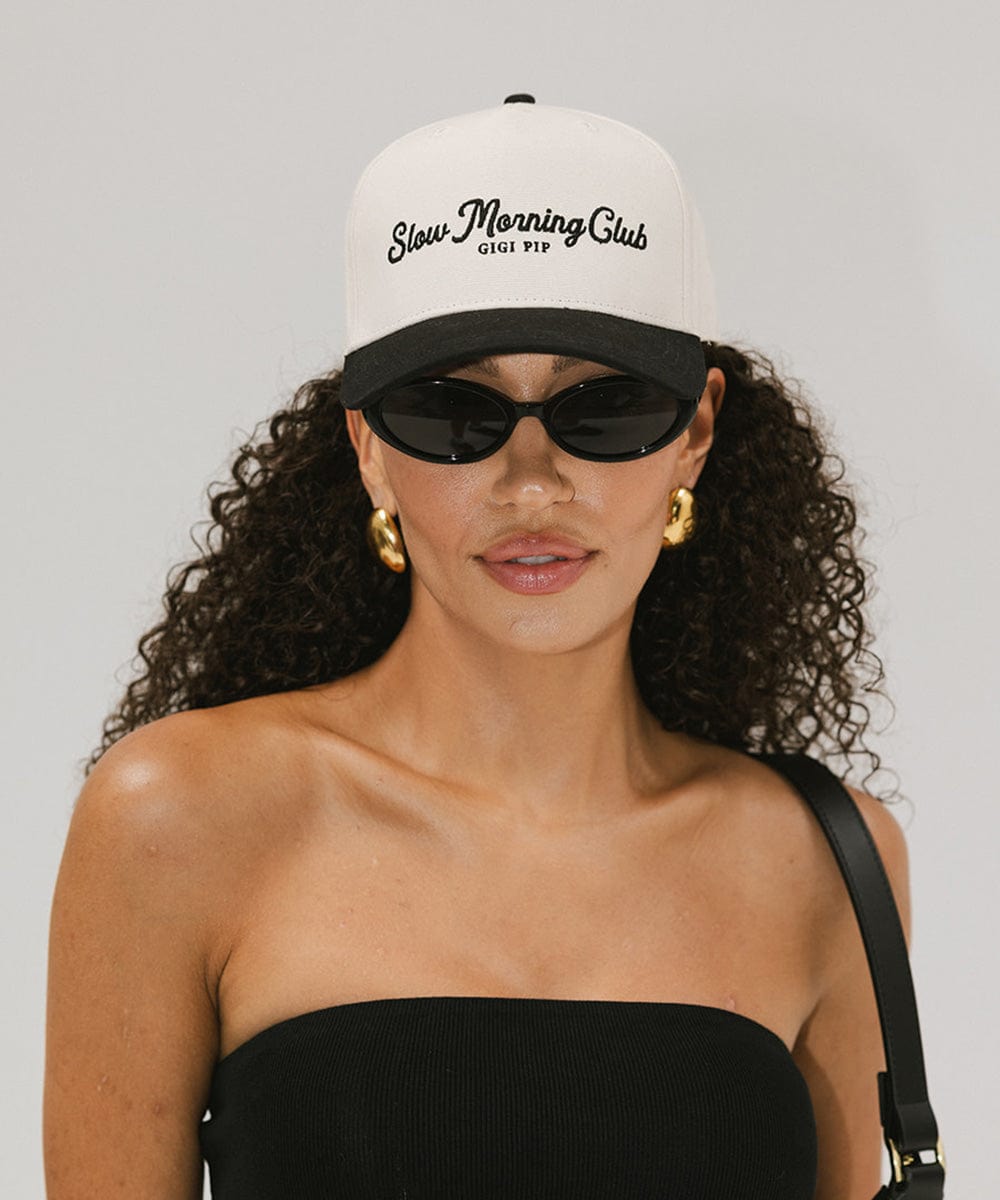Woman wearing a cream and black cap with the text "Slow Morning Club", sunglasses, and a black top against a plain background #color_cream - black