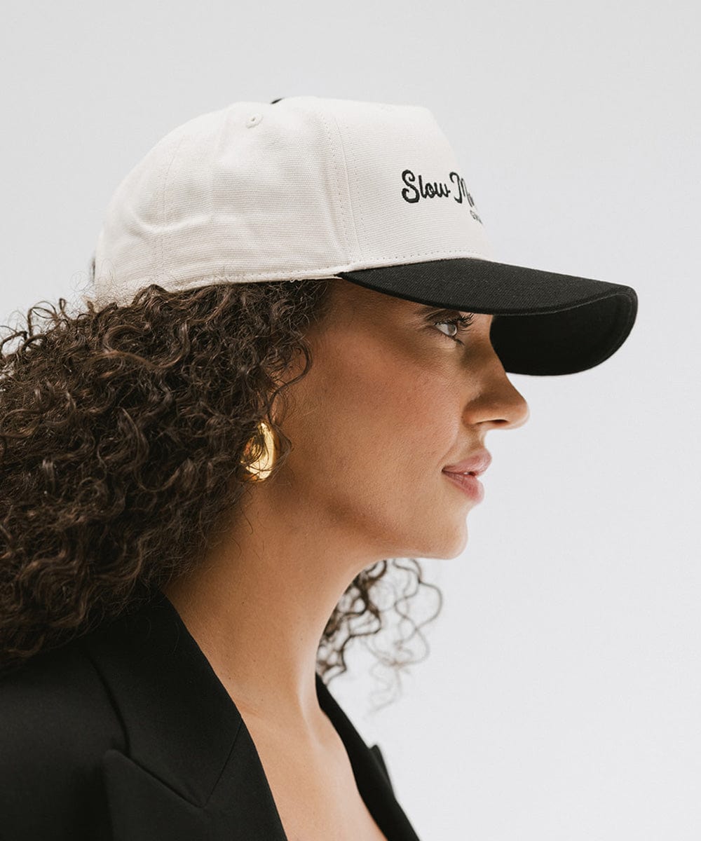 Woman wearing a cream and black cap with the text "Slow Morning Club", and a black coat, facing sideways against a plain background #color_cream - black