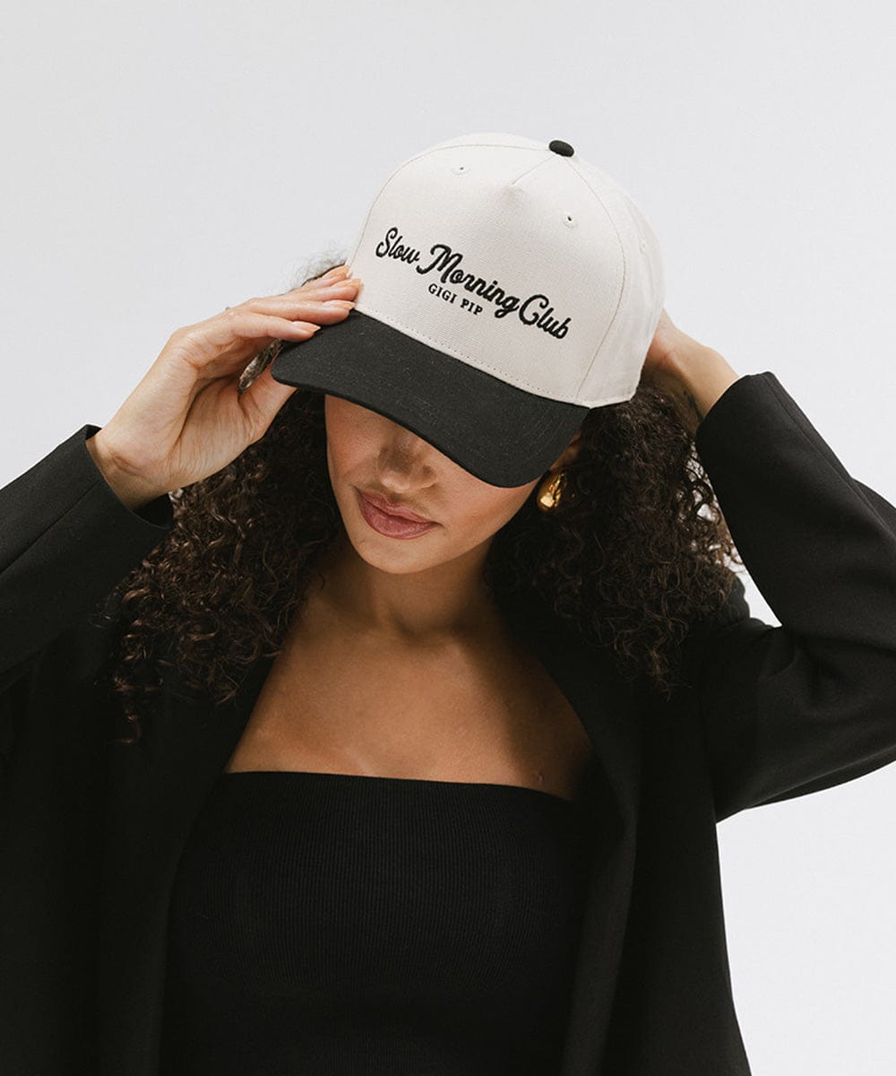 Woman wearing a cream and black cap with the text "Slow Morning Club", and a black top with a black coat, against a plain background #color_cream - black