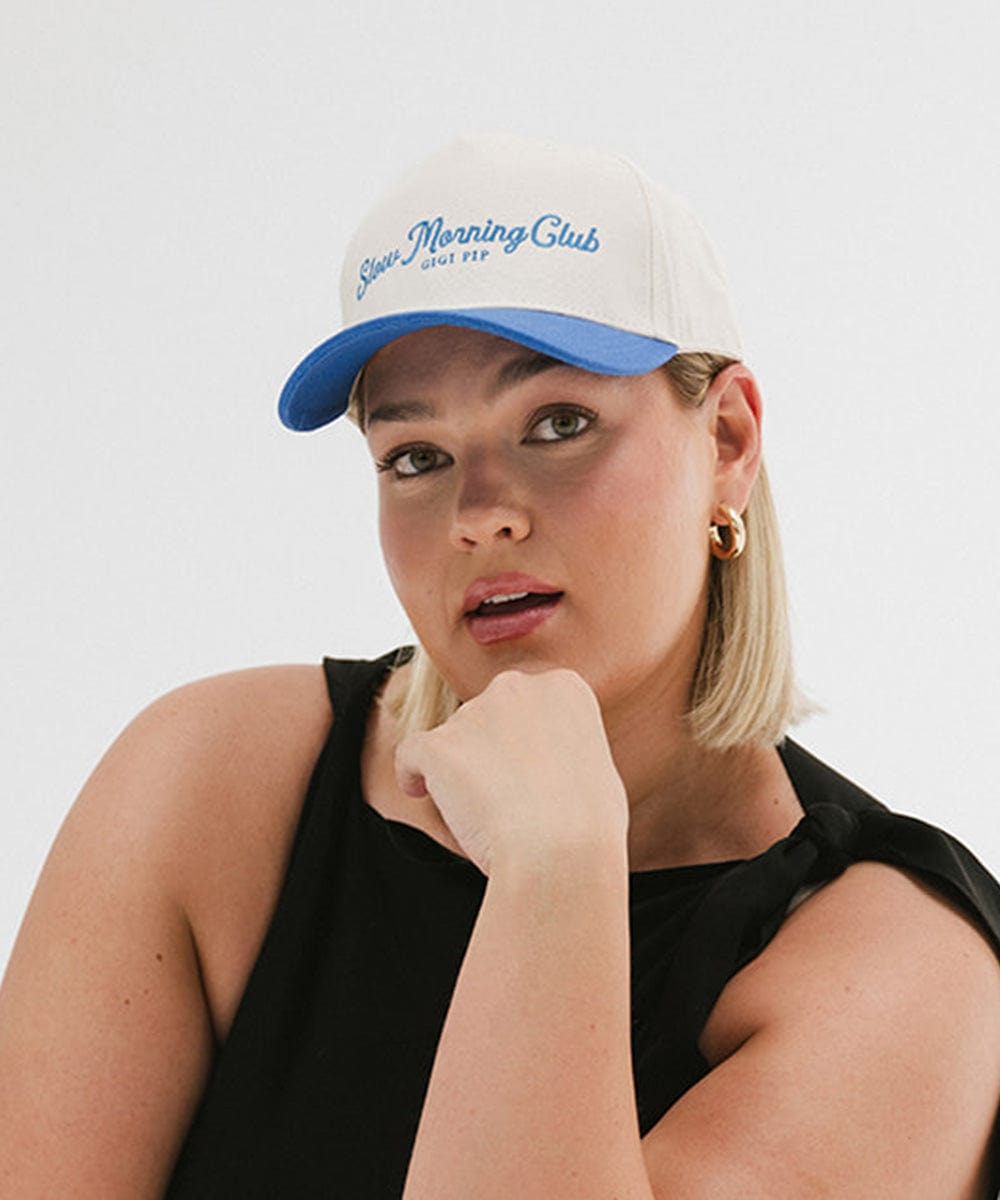 Woman wearing a cream and blue cap with 'Slow Morning Club' text and a black top against a plain background #color_cream - blue