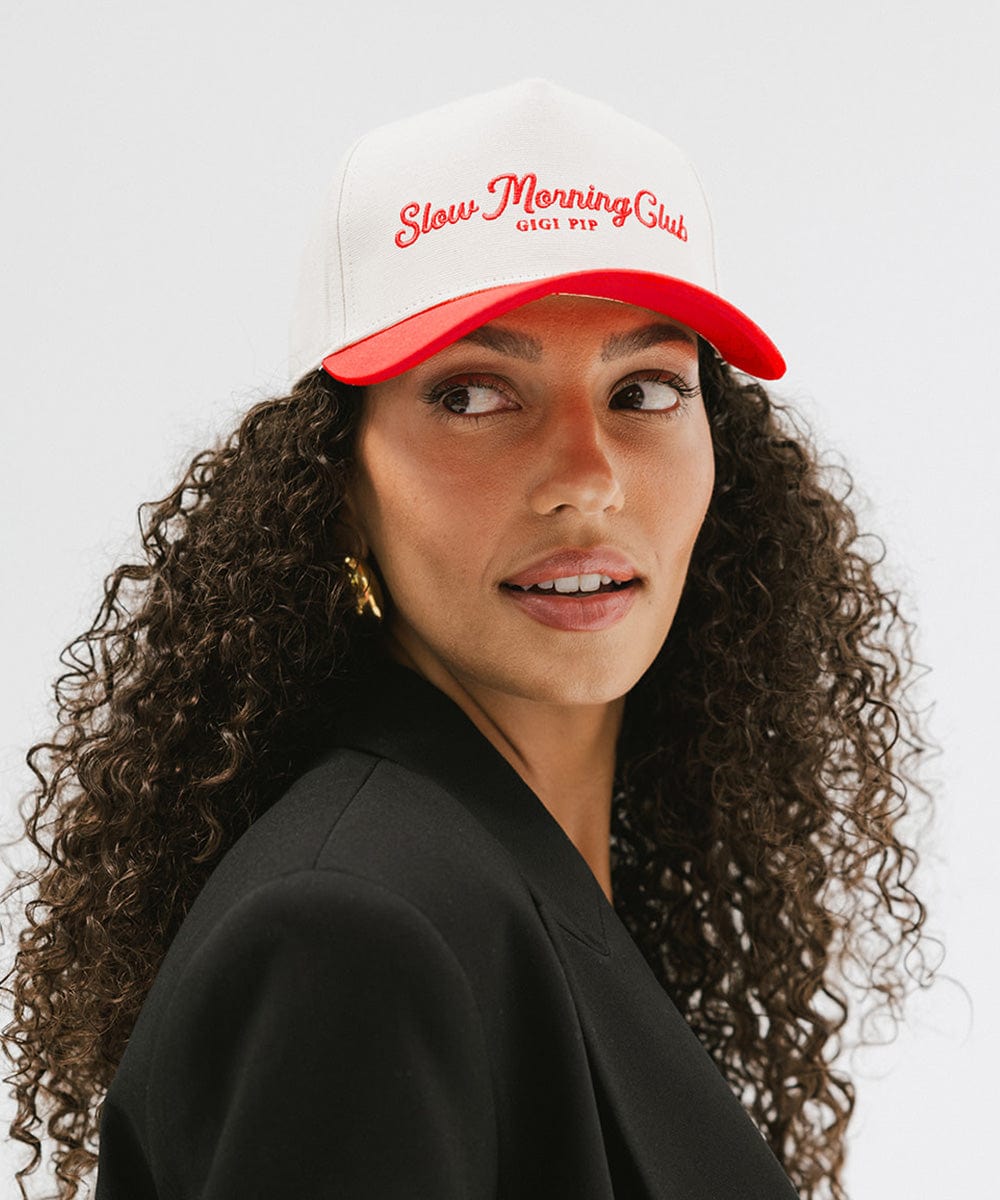 Woman with curly hair wearing a cream and red cap with 'Slow Morning Club' text and a black coat against a plain background #color_cream - red