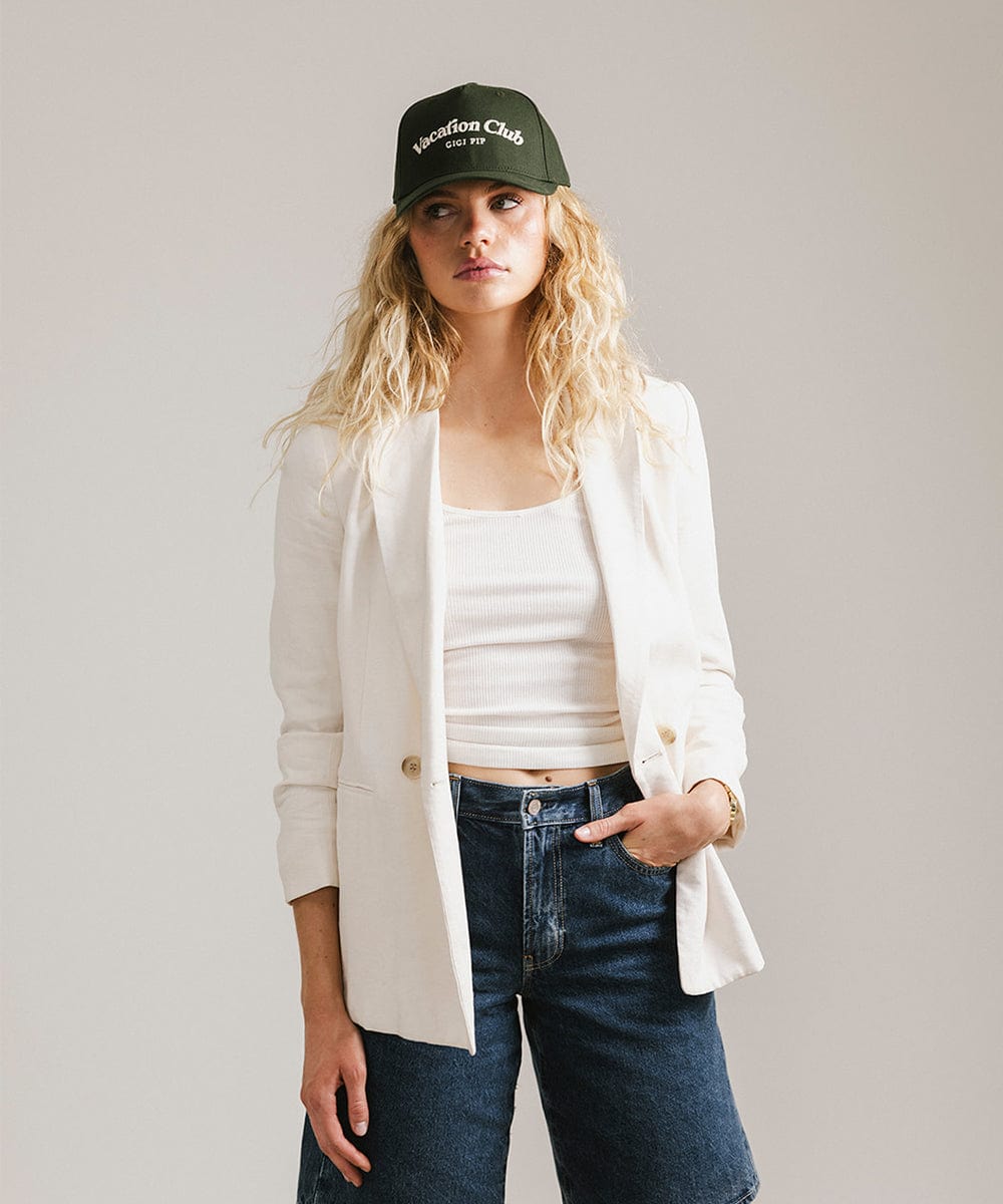 Woman wearing a dark green cap with 'Vacation Club' text, a white top with a white coat and denim shorts against a plain background #color_dark green