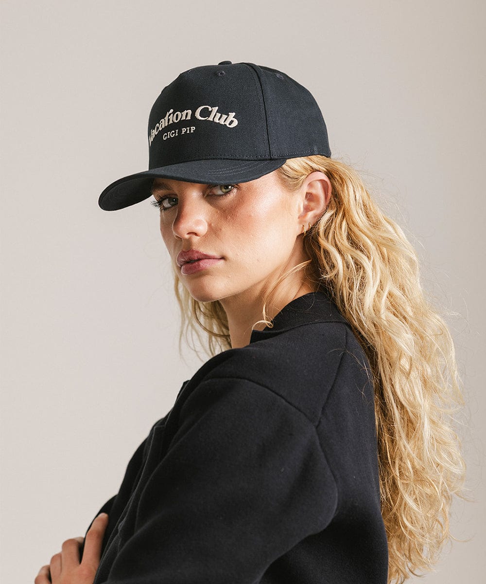 Woman wearing a navy cap with 'Vacation Club' text and a black shirt, posing sideways against a plain background #color_navy