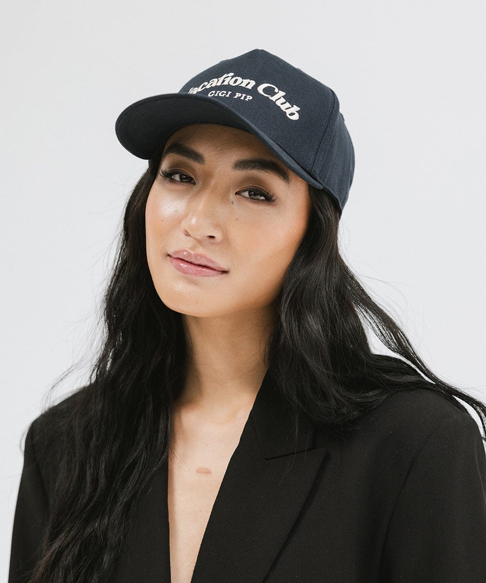 Woman wearing a navy cap with 'Vacation Club' text and a black blazer against a plain background #color_navy