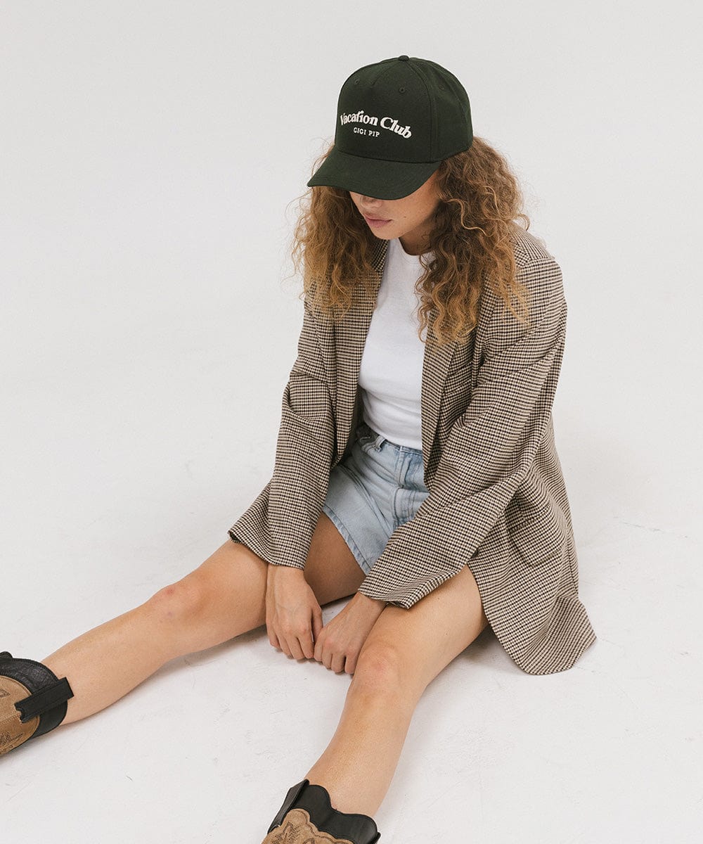 Woman wearing a dark green cap with 'Vacation Club' text, a white top with a coat and denim shorts, sitting against a plain background #color_dark green
