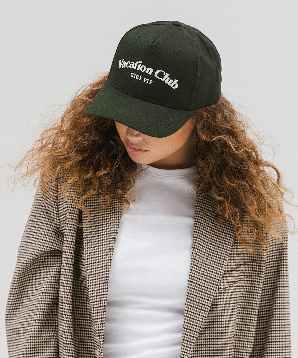 Woman wearing a dark green cap with 'Vacation Club' text and a white top with a coat against a plain background #color_dark green