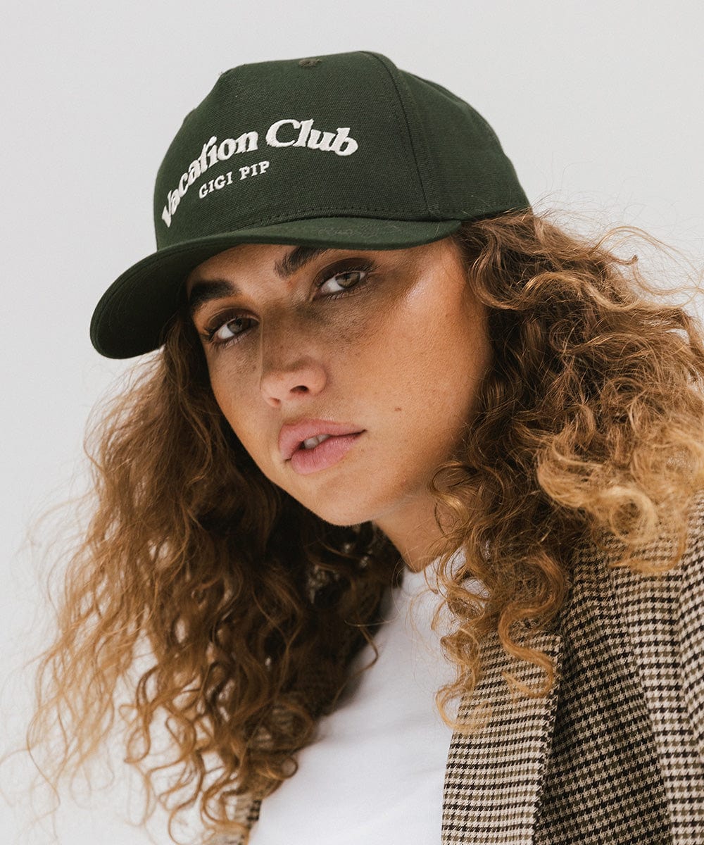 Woman wearing a dark green cap with 'Vacation Club' text and a white top against a plain background #color_dark green