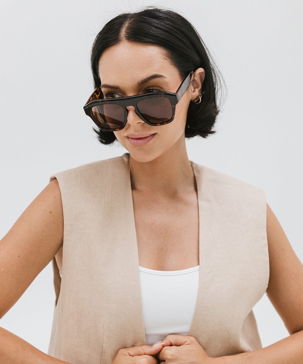 Woman wearing tortoise colored sunglasses and a white top with a beige jacket against a plain background #color_tortoise