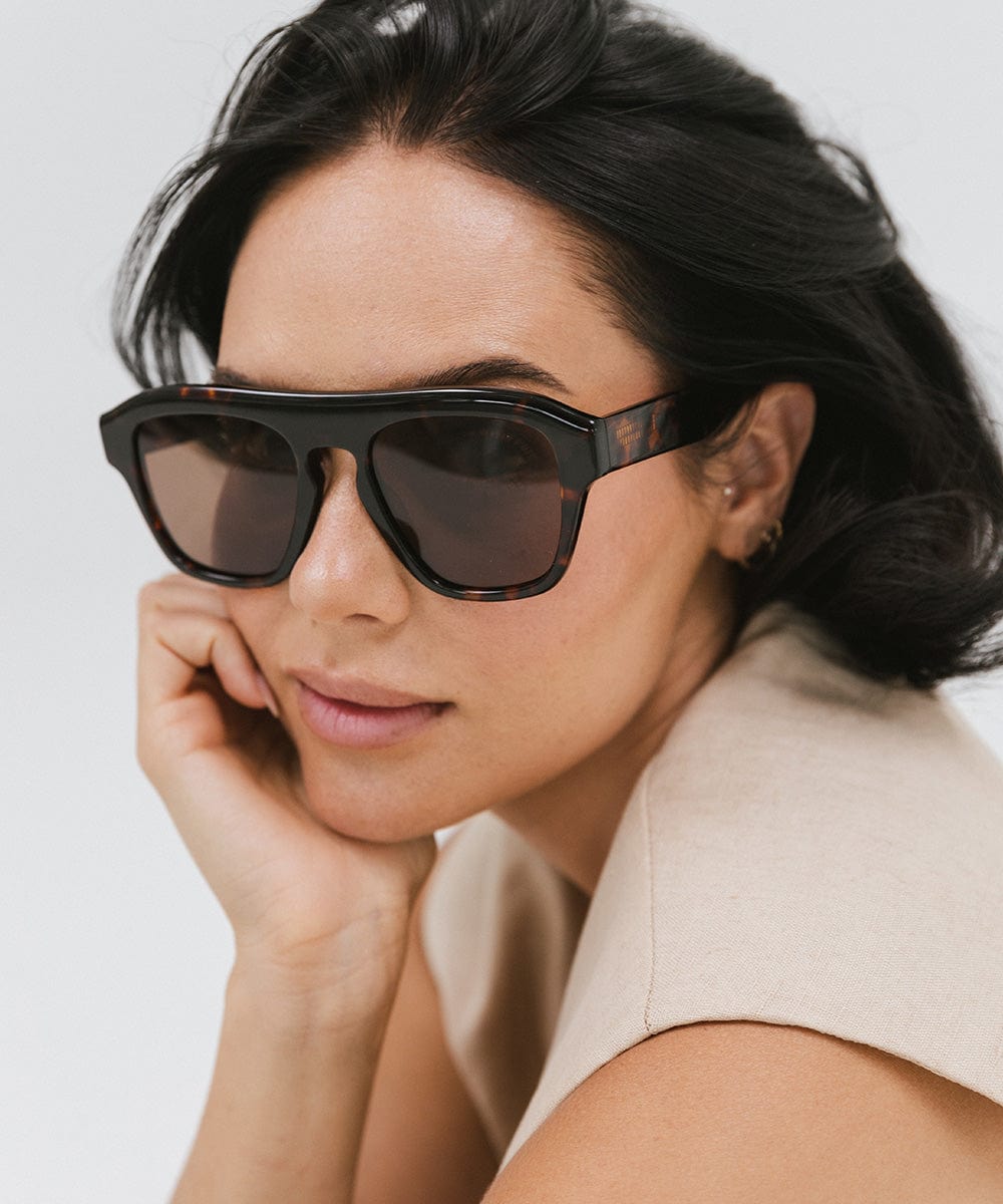 Woman wearing tortoise colored sunglasses and a beige top, posing against a plain background #color_tortoise