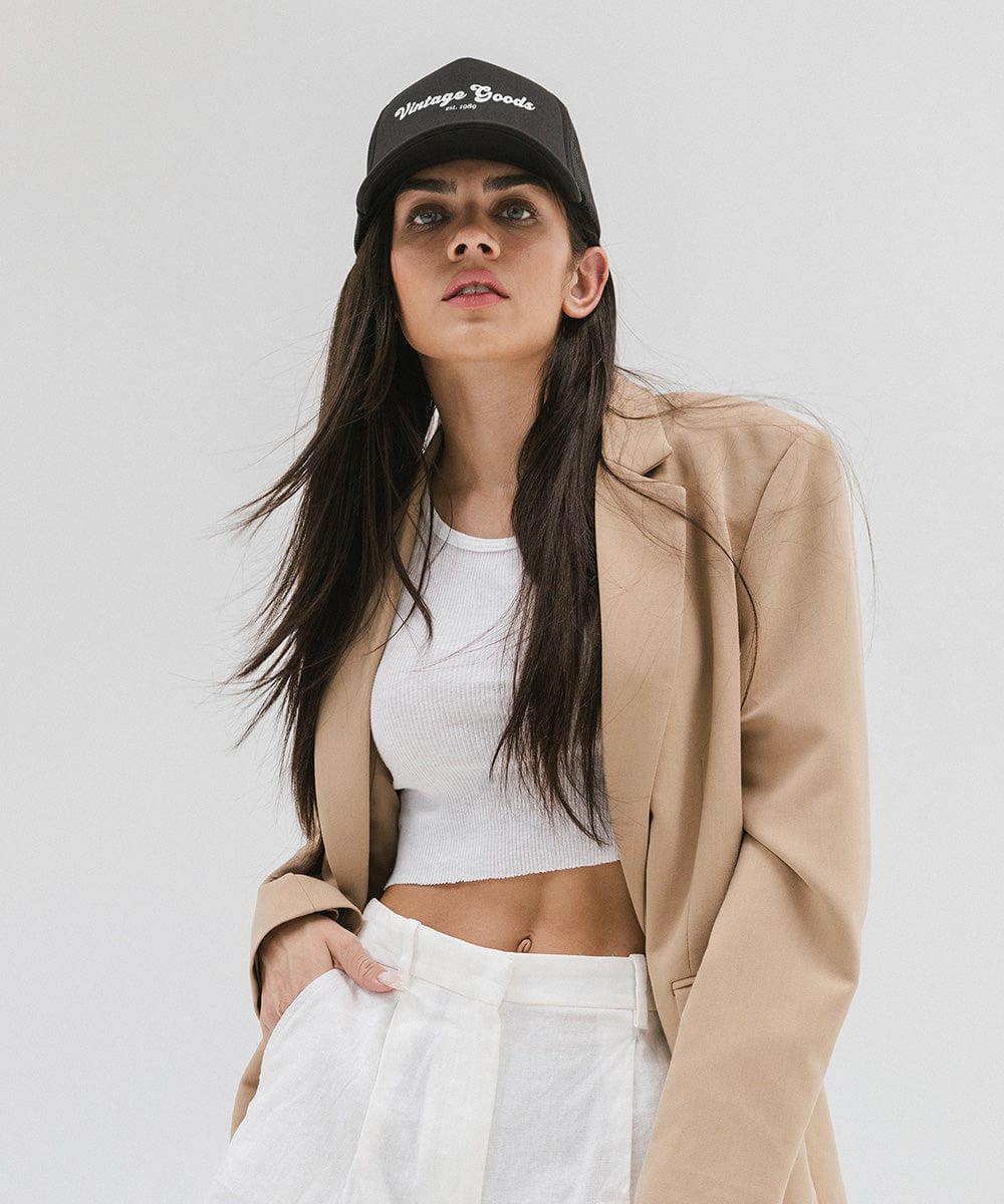 Woman wearing a black trucker hat with 'Vintage Goods' text, a white top with a beige blazer and white pants against a plain background #color_black