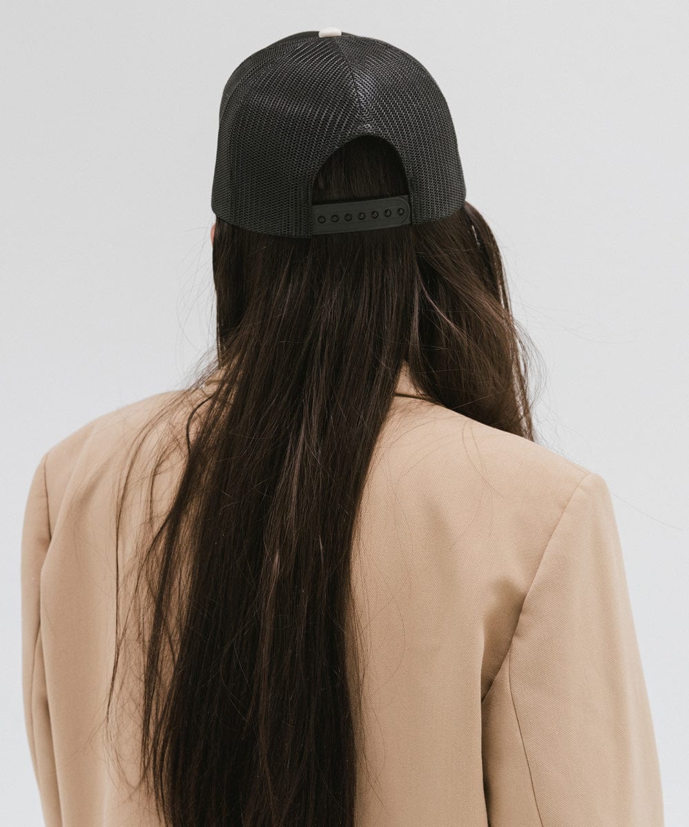 Woman wearing a black trucker hat with 'Vintage Goods' text and a beige blazer, facing behind against a plain background #color_black