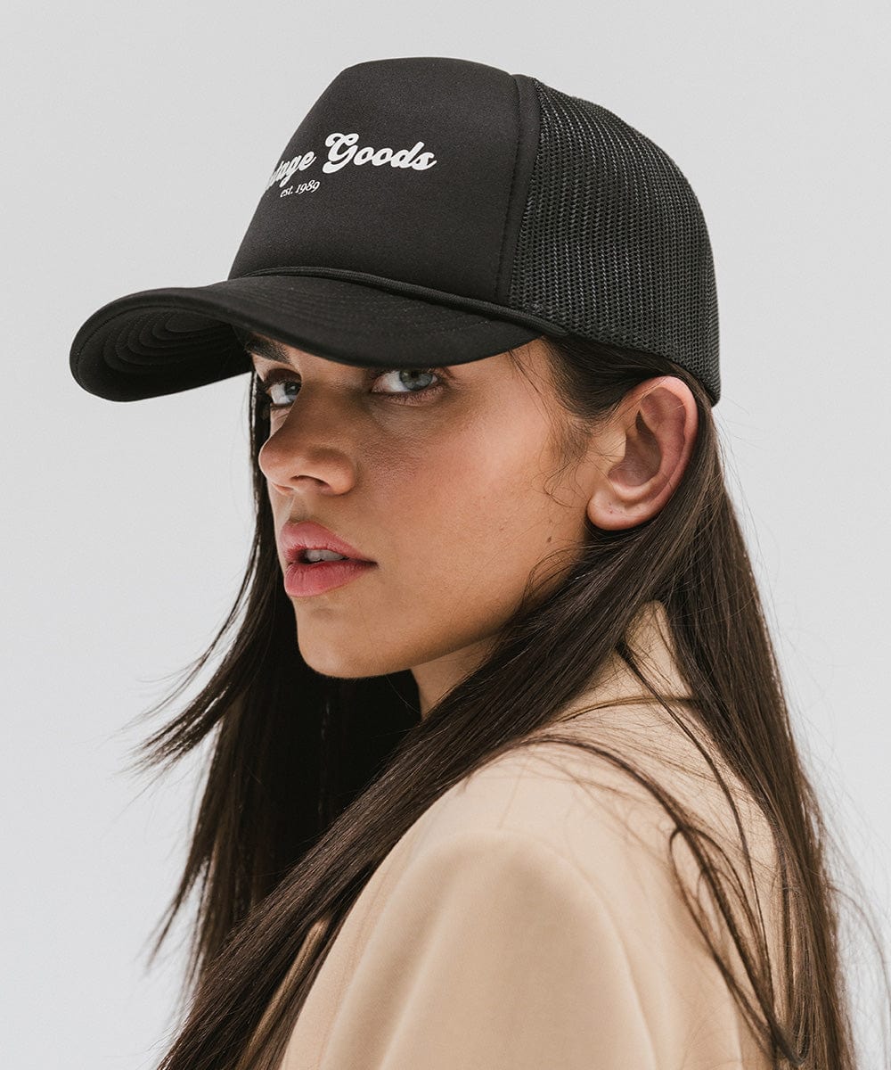 Woman wearing a black trucker hat with 'Vintage Goods' text and a beige blazer against a plain background #color_black