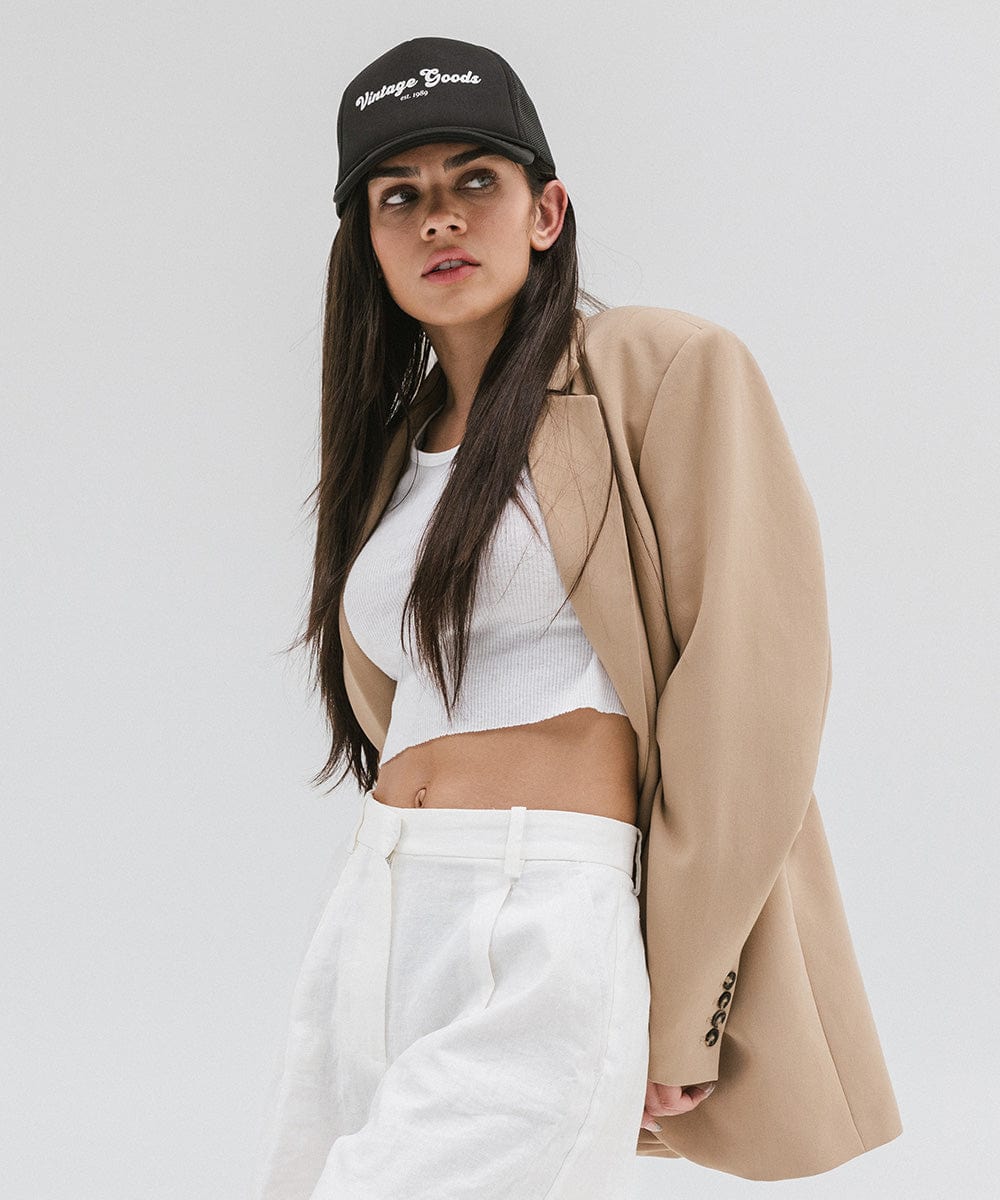 Woman wearing a black trucker hat with 'Vintage Goods' text, a white top with a beige blazer and white pants, looking sideways against a plain background #color_black