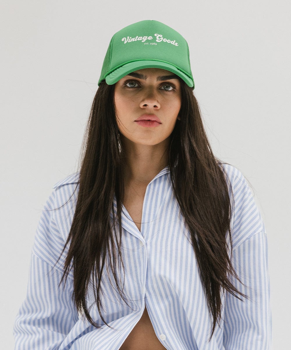 Woman wearing a vintage green colored trucker hat with 'Vintage Goods' text and a white shirt with blue lines against a plain background #color_vintage green