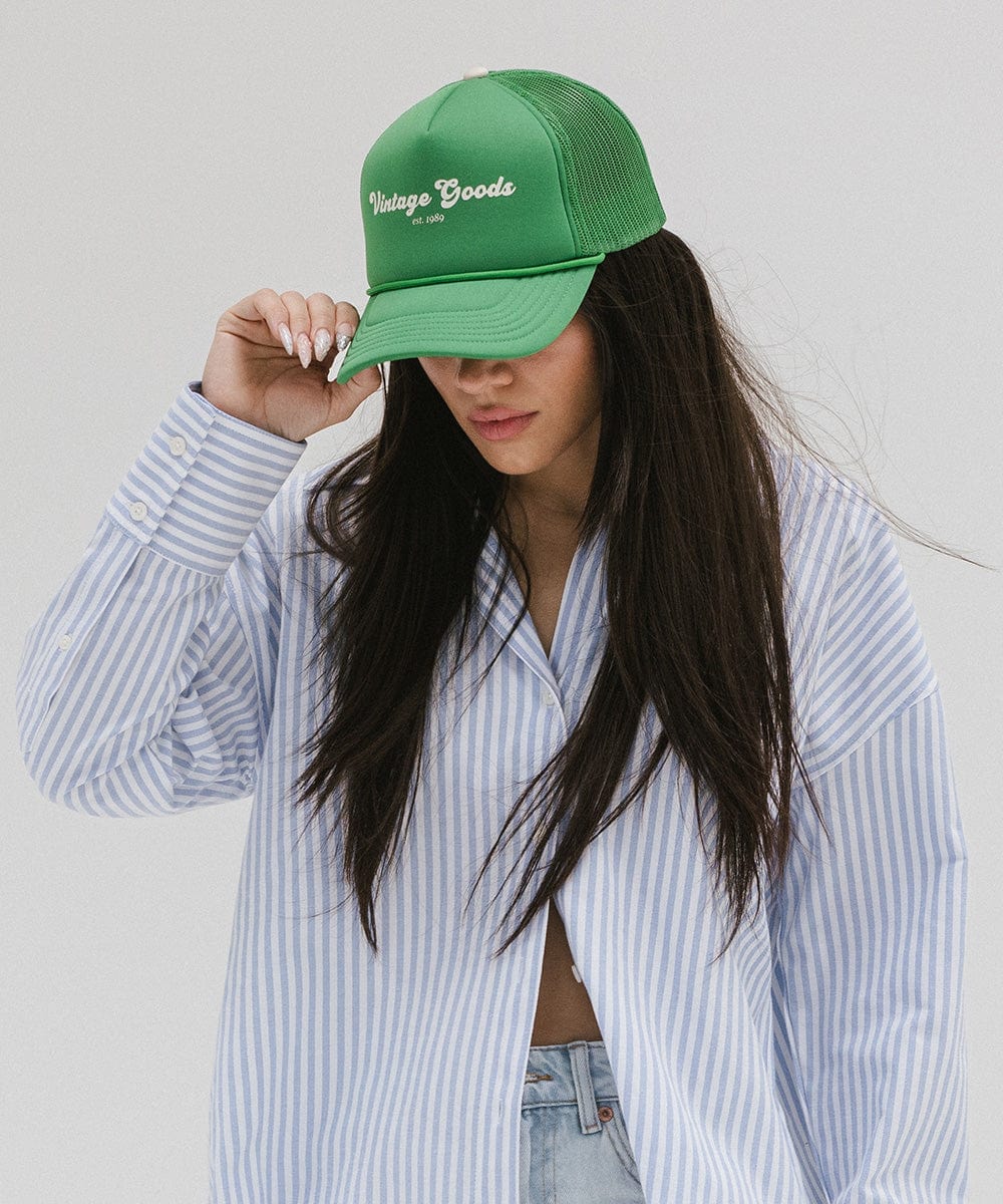 Woman wearing a vintage green colored trucker hat with 'Vintage Goods' text and a white shirt with blue lines, posing against a plain background #color_vintage green